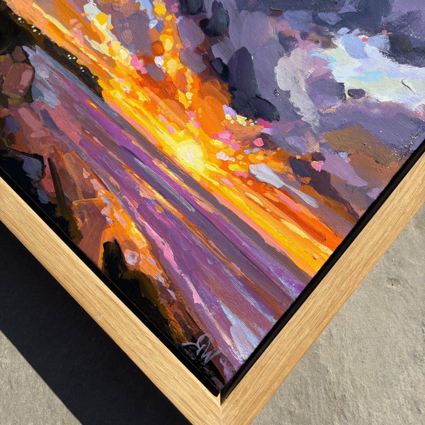 A framed abstract painting featuring a dramatic diagonal composition with a bright yellow-orange burst surrounded by purple, pink, and gray brushstrokes, set in a light wood frame on a concrete surface.