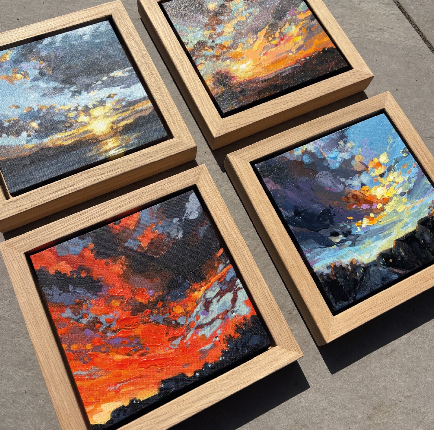 Four small paintings of sunset scenes in wooden frames on a concrete surface.