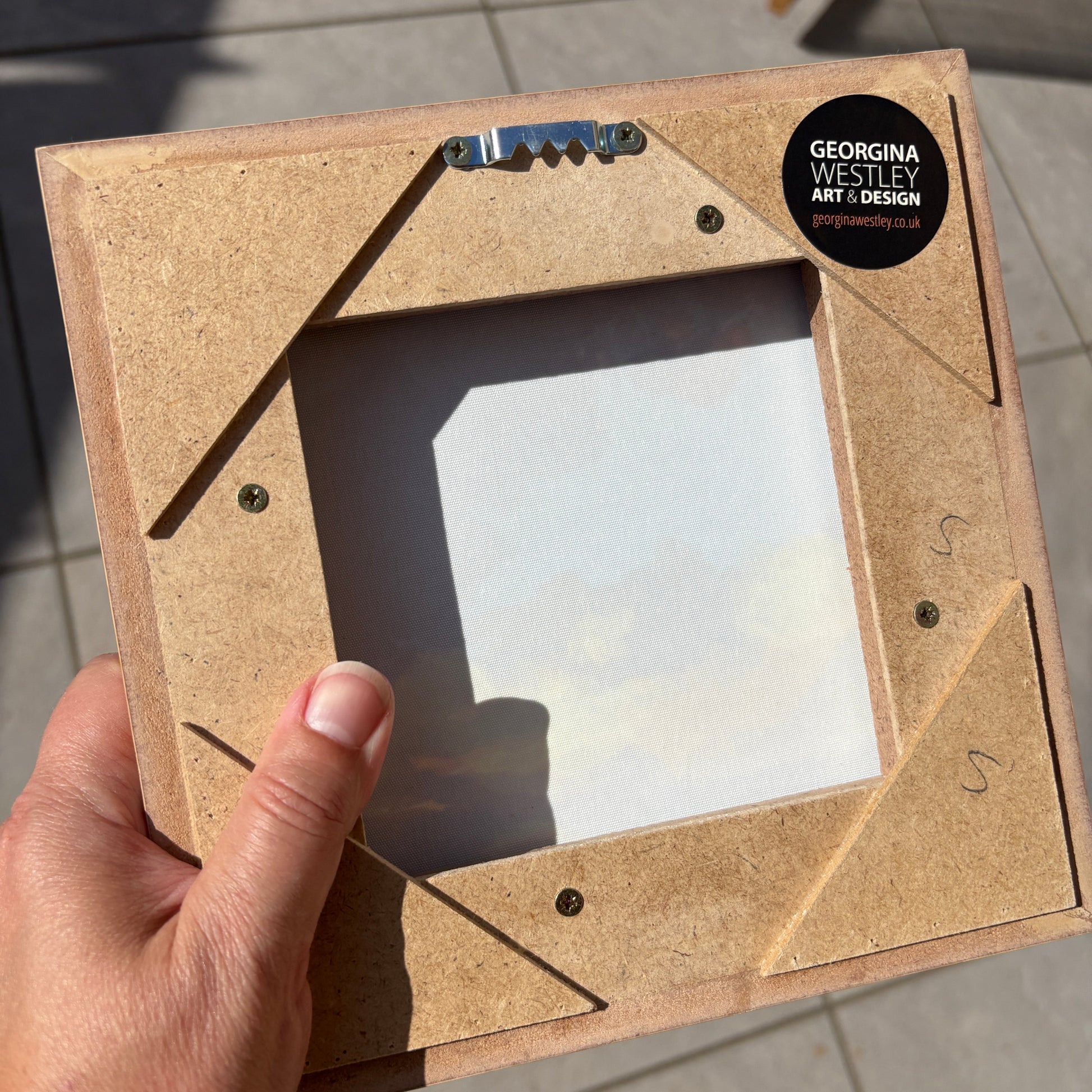 A hand shows the back of the frame, with a sawtooth hanger and sticker that reads 'GEORGINA WESTLEY ART DESIGN'. The background shows outdoor paving stones in bright sunlight.