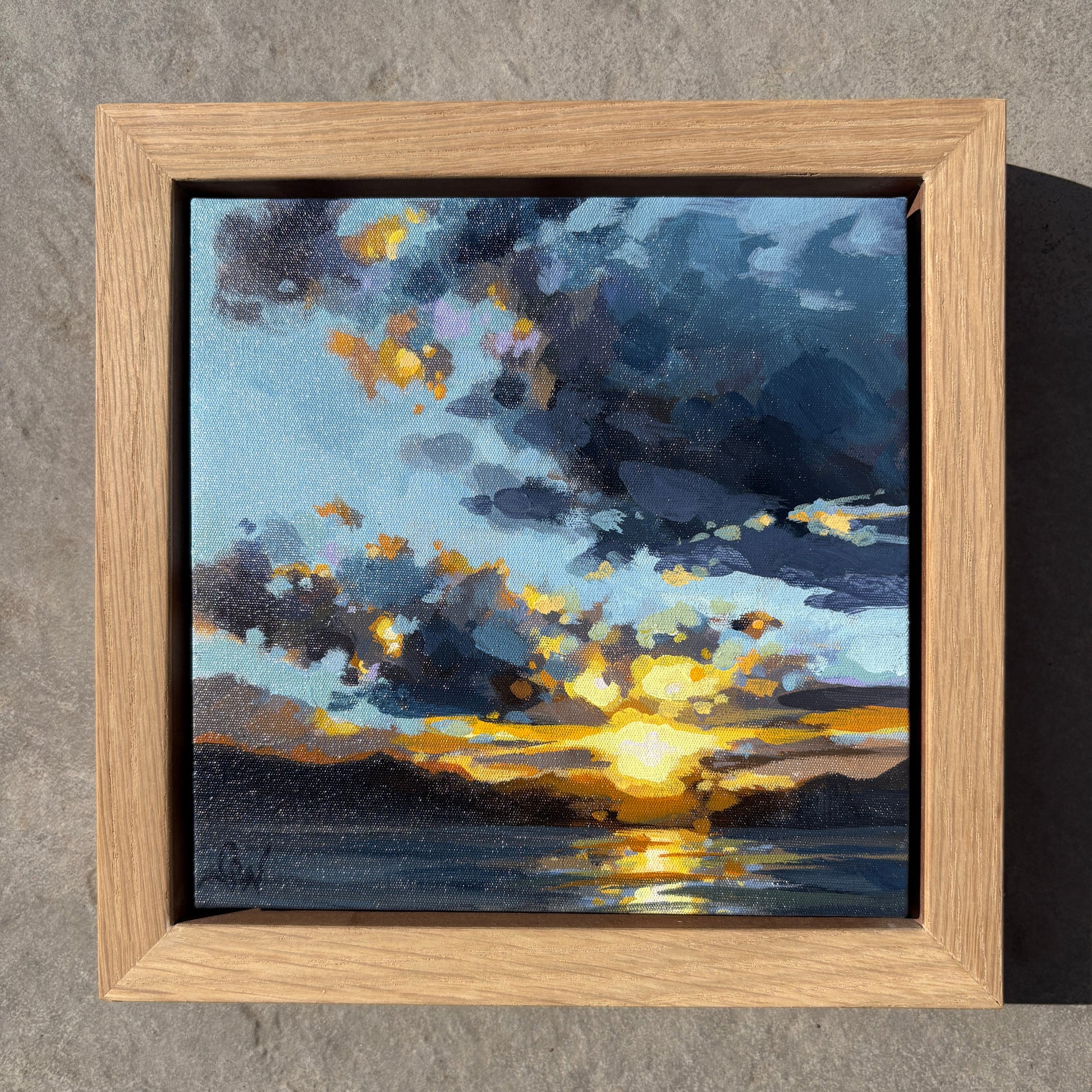 A square framed painting of a dramatic sunset over water, featuring dark blue and gray clouds with bright yellow sunlight reflecting on the horizon, set in a light oak wood frame against a textured gray concrete background.