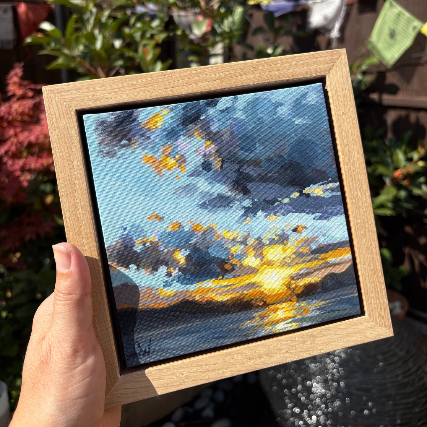 A hand holds a square framed painting of a dramatic sunset over water, with blue and gray clouds and bright yellow light reflecting on the horizon. The painting is set in a natural light oak frame, photographed outdoors against a blurred garden background.