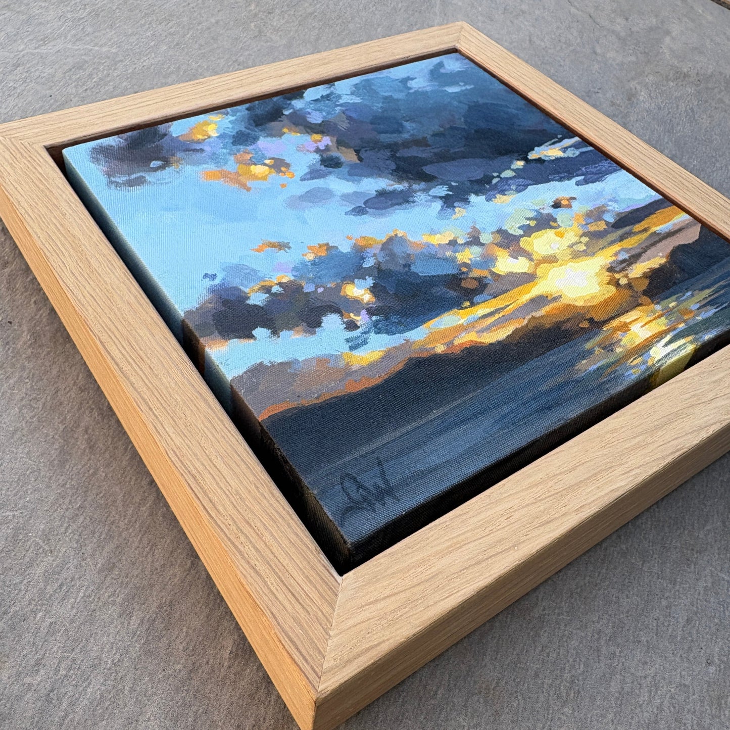 A framed square canvas painting of a dramatic sunset sky with blue, gray, and golden-yellow clouds, displayed on a gray stone surface.
