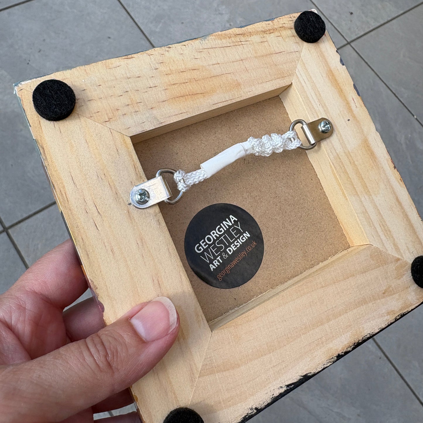 A hand holding a square wooden picture frame with a natural light wood finish, black velvet corners, and a clear beaded hanging loop on the back. A circular sticker on the backing reads 'GEORGINA Westley ART DESIGN'.