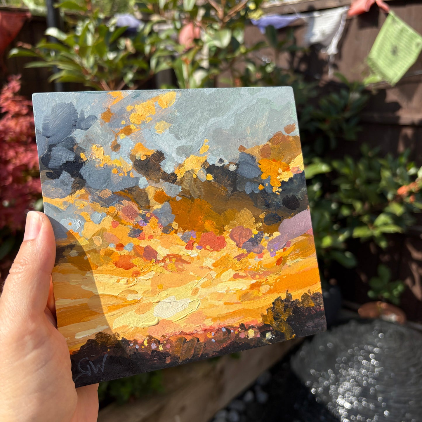 A hand holds a small square canvas painting outdoors in a garden setting. The painting features an abstract landscape with a bright orange and yellow sky, gray clouds, and dark tree silhouettes at the bottom.