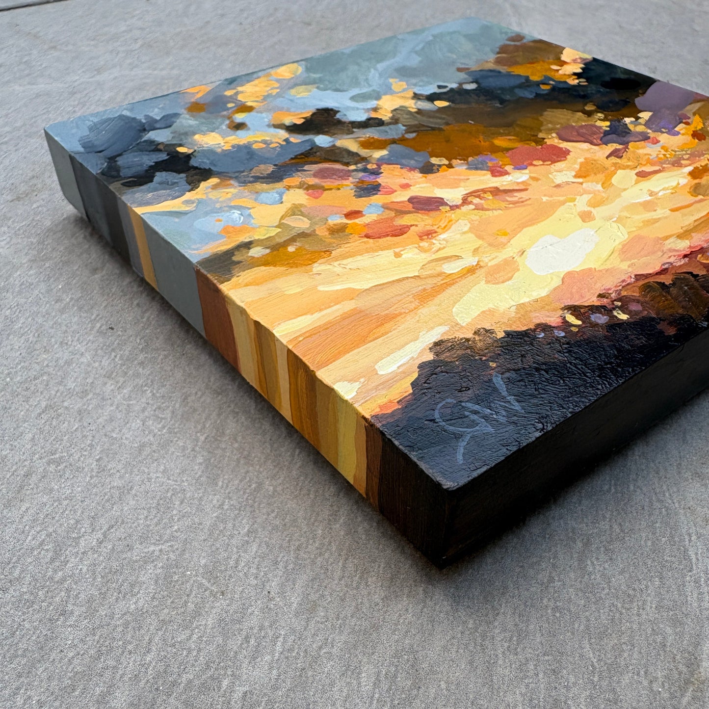 An abstract rectangular canvas painting with thick impasto brushstrokes in black, gold, orange, yellow, blue-gray, and brown, photographed at an angle on a textured gray surface.