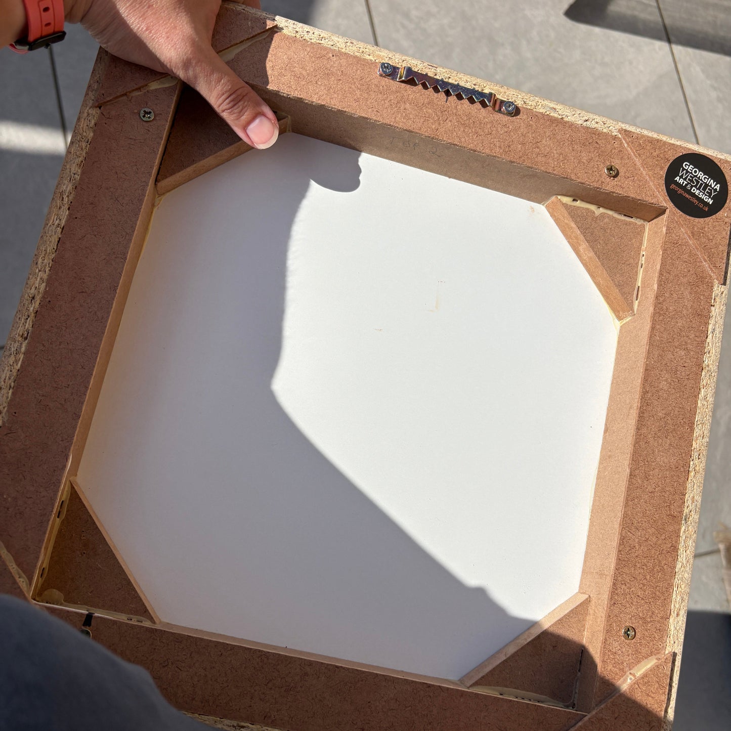 A person holding a square picture frame with a white canvas insert, showing the back construction of the frame. The frame has a natural brown wood/mahogany finish and a black circular sticker on the top right corner.