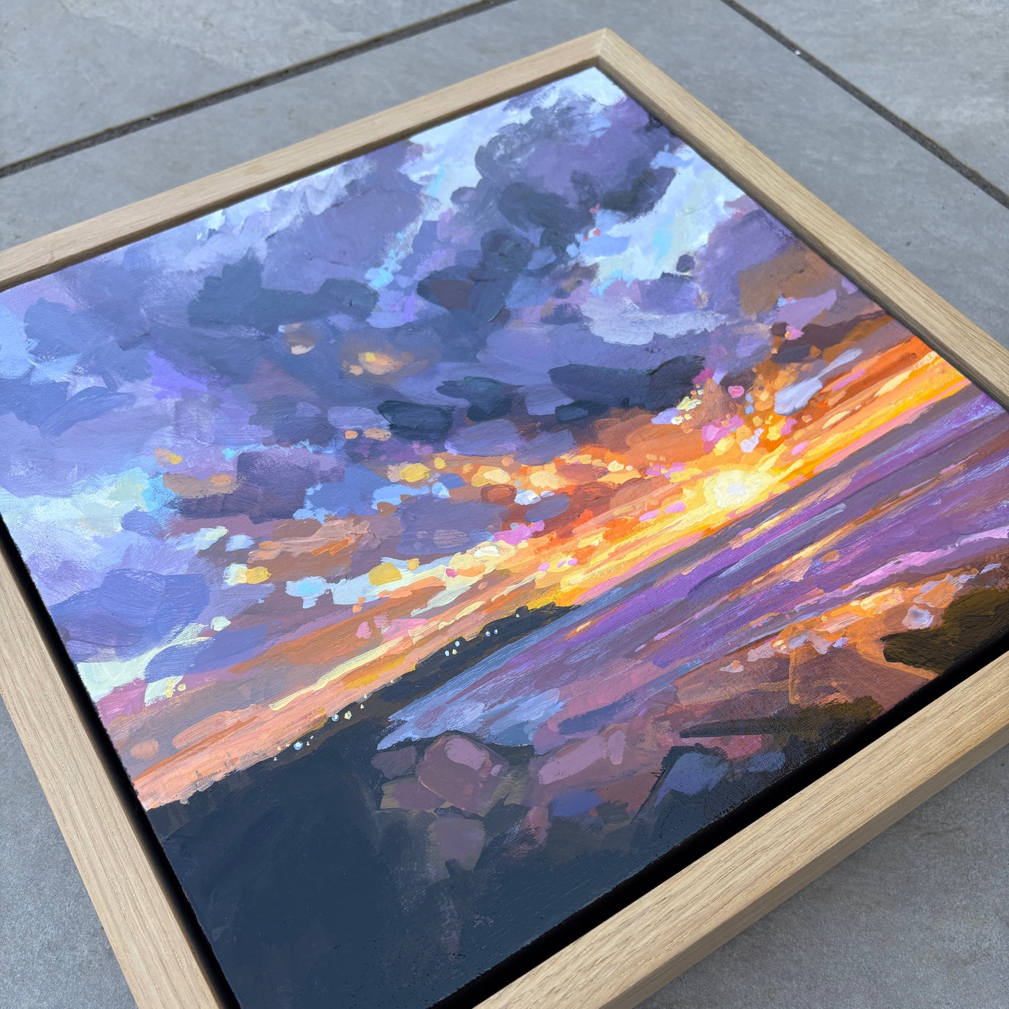 A square framed canvas painting of a dramatic sunset sky, featuring purple, blue, orange, and yellow brushstrokes, set in a light natural wood frame on a gray tiled floor.