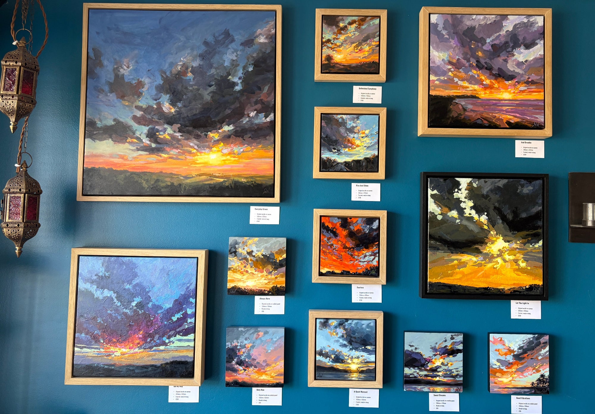 A gallery-style wall display featuring multiple framed landscape paintings of stormy sunsets and clouds, mounted on a deep blue wall with small white labels beneath each piece.