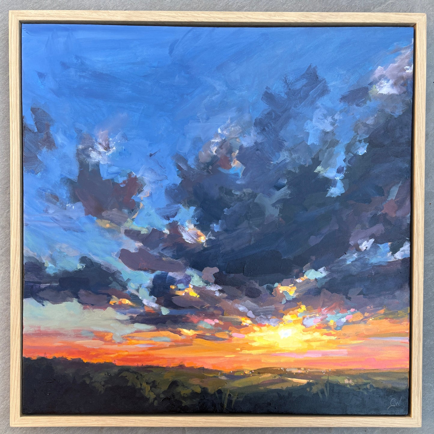 A framed landscape painting of a dramatic sunset sky with dark blue and purple clouds, a bright yellow sun near the horizon, and orange light over dark green hills, displayed on a gray tiled floor with shoes visible at the bottom.