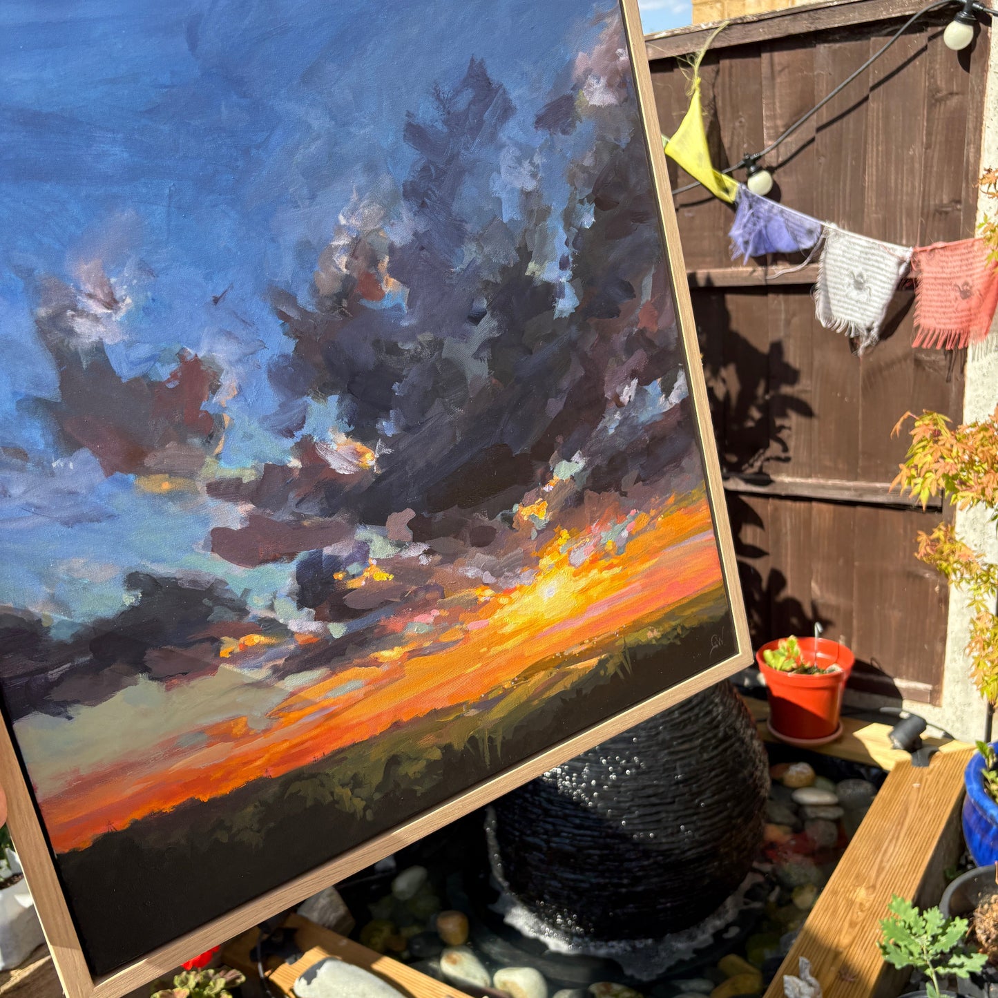 A large framed landscape painting of a dramatic sunset with dark storm clouds and a glowing orange horizon, displayed in a backyard garden setting.