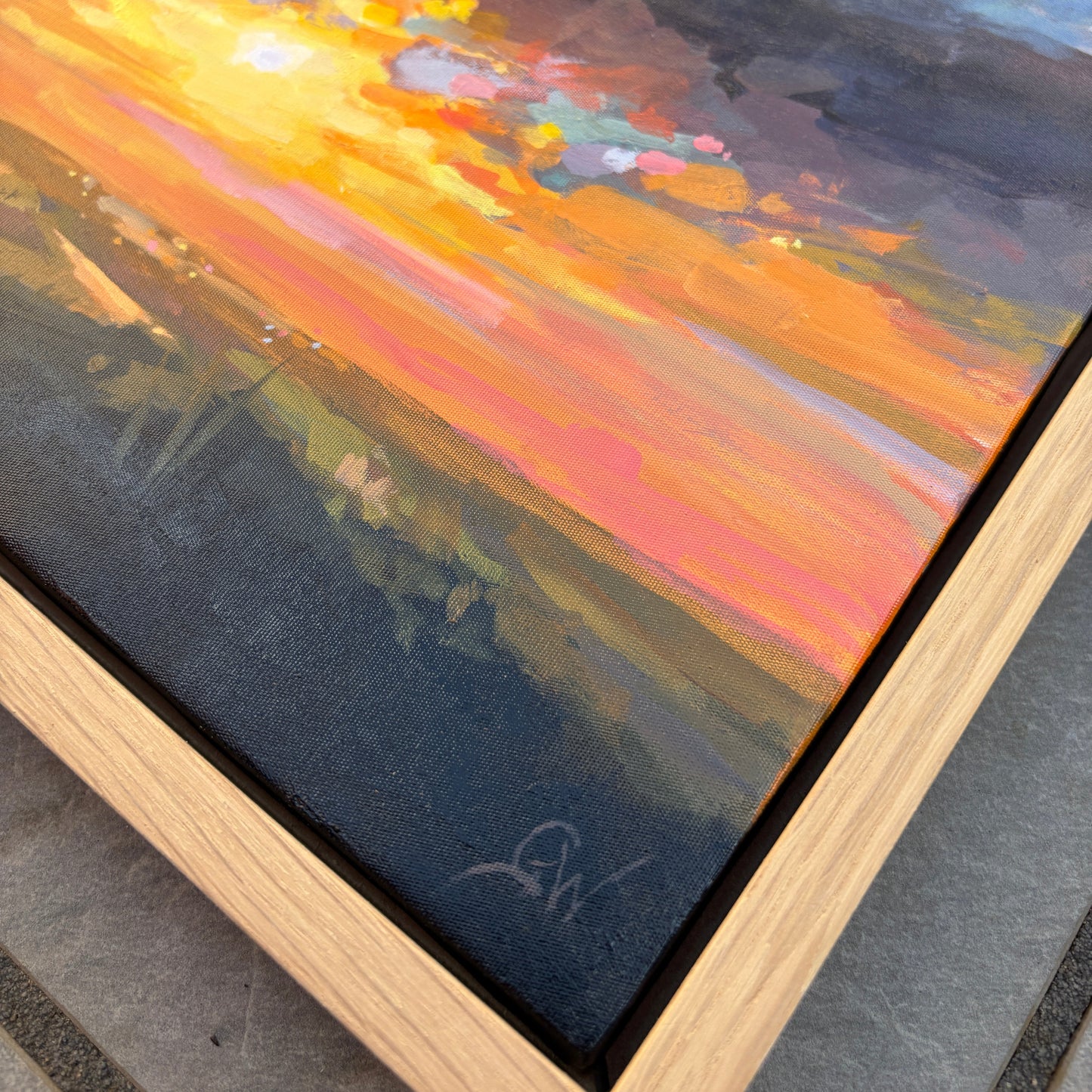 A framed landscape oil painting showing a dramatic sunset over a field, with a light oak wood frame. The painting features vibrant orange, yellow, pink, and blue brushstrokes, and a signature is visible in the bottom right corner.