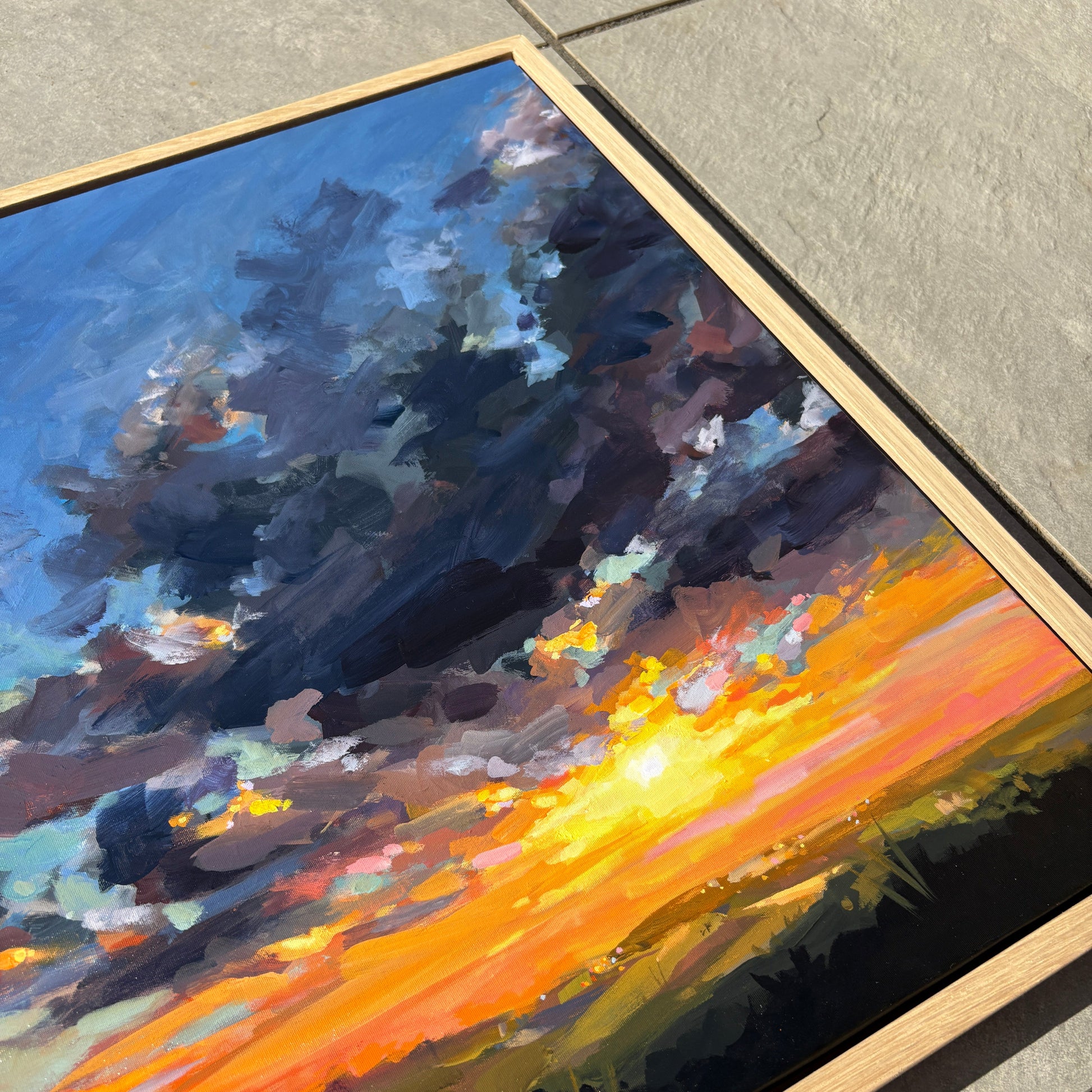 A framed landscape oil painting depicting a dramatic sunset over a field, resting on a gray tiled floor. The painting features a bright yellow and orange sunburst, dark blue and purple storm clouds, and green fields under a blue sky, housed in a light natural wood frame.