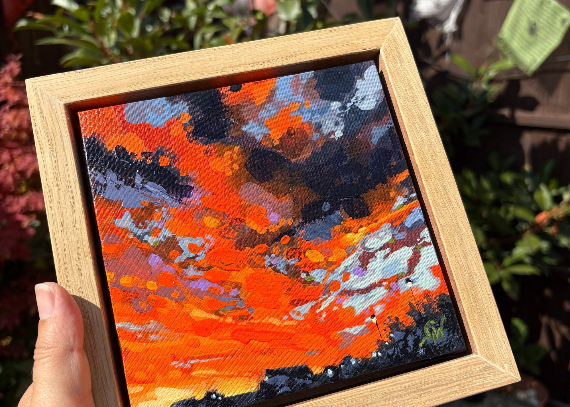 A hand holds a square wooden frame containing an expressive acrylic painting of a fiery orange and red sky with dark clouds, set against a blurred outdoor garden background.