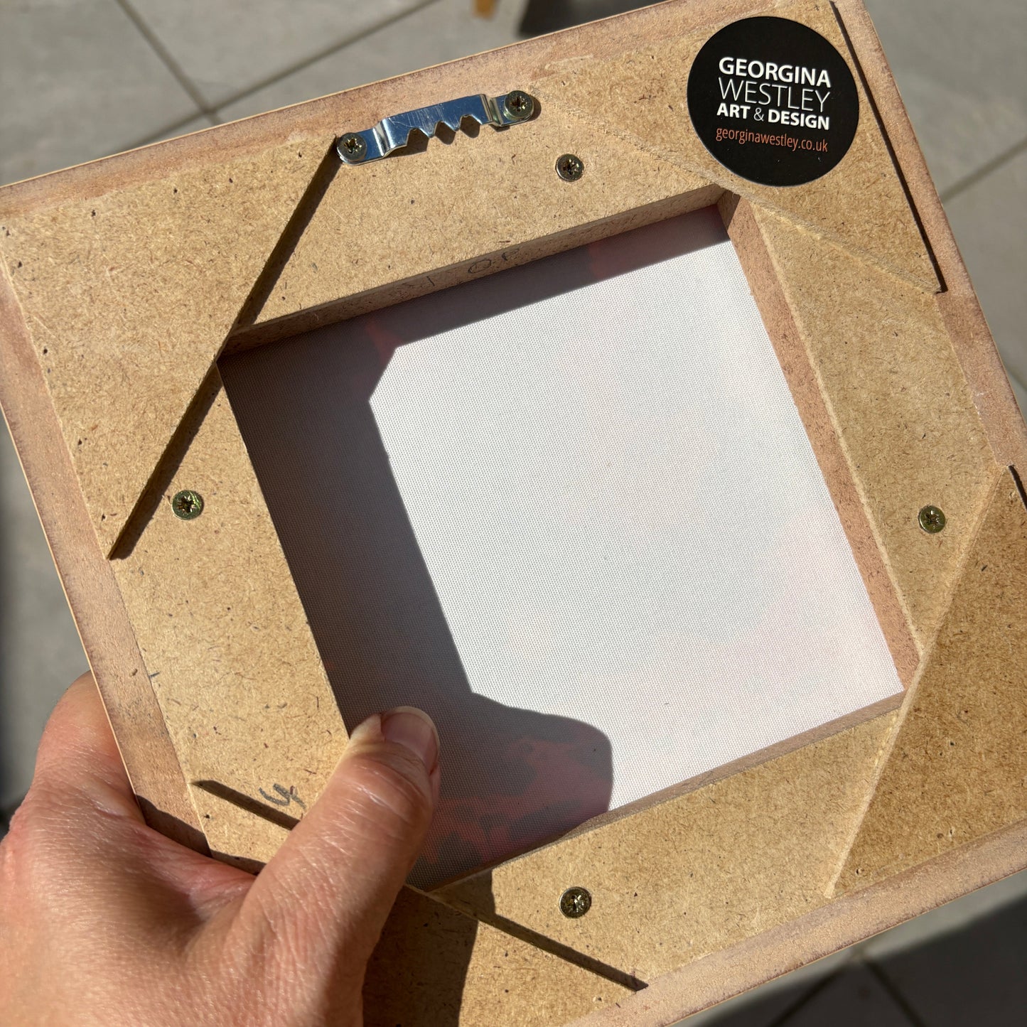 A hand holding a square picture frame made from natural particle board with a clear glass or plastic insert, photographed outdoors on a tiled floor. A black circular sticker on the frame reads 'GEORGINA WESTLEY ART & DESIGN'