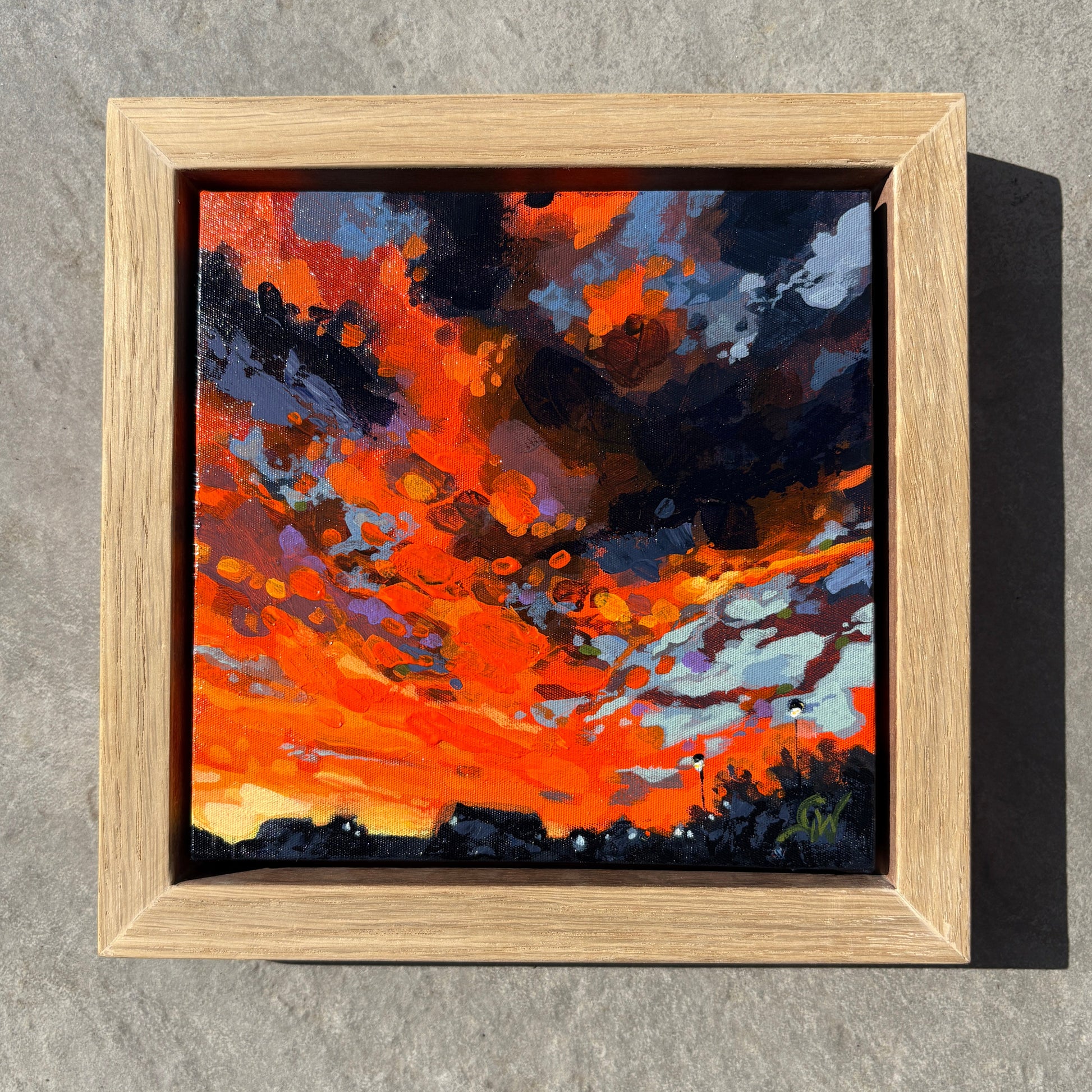 A square framed painting of a dramatic orange and red sunset sky with dark clouds, displayed on a textured gray concrete wall.