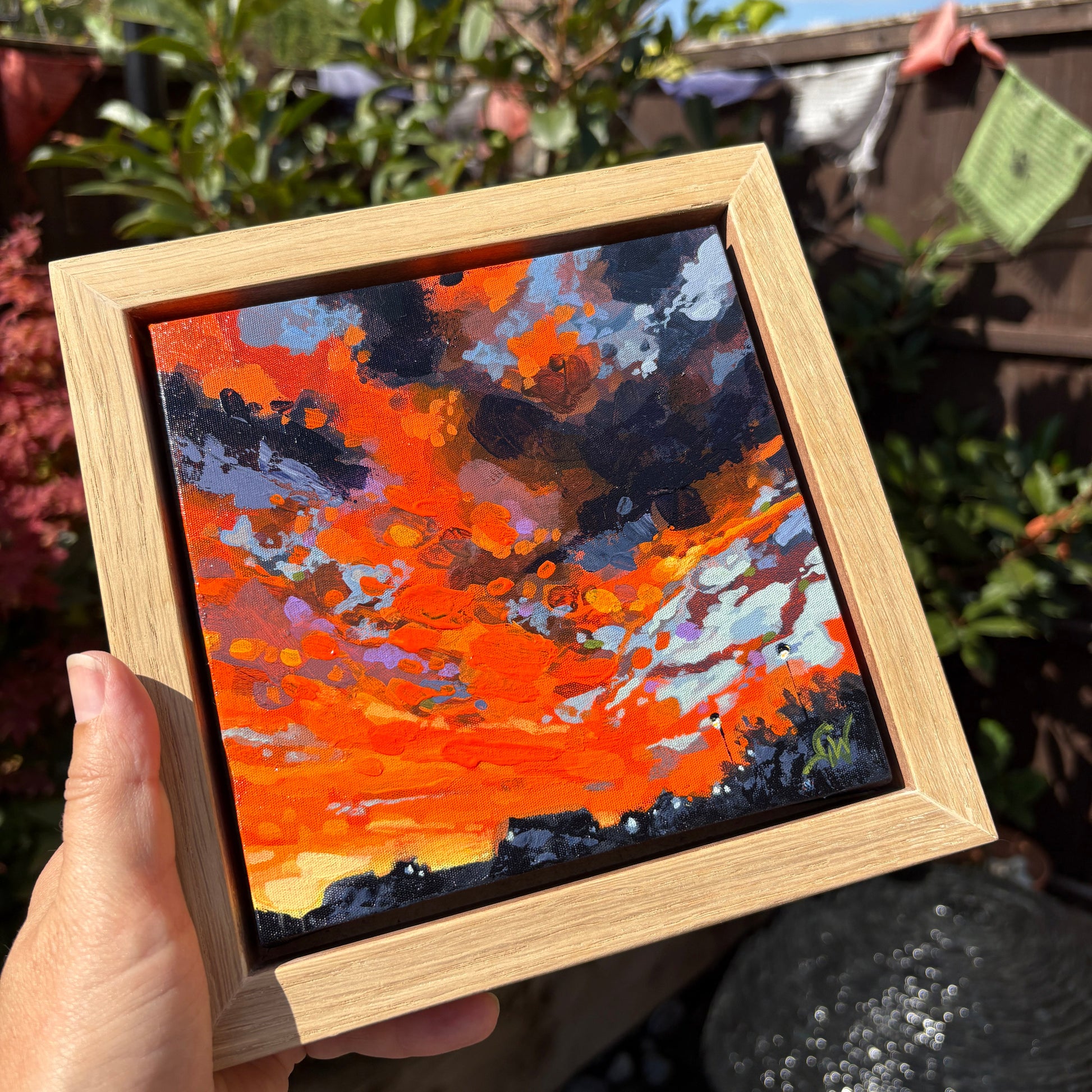 A hand holds a square wooden frame containing an expressive oil painting of a fiery orange and red sky with dark clouds, set against a blurred outdoor garden background.