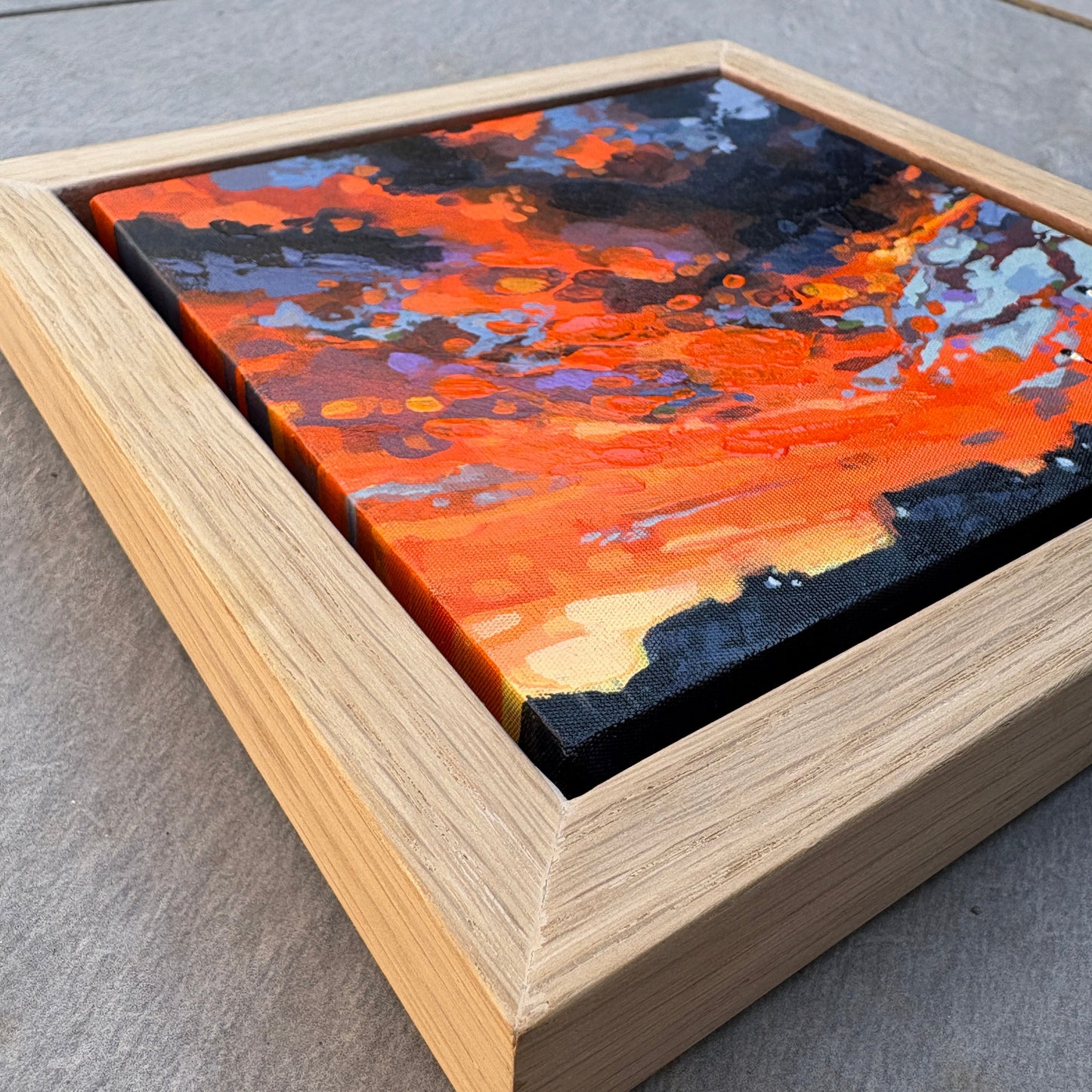 A framed abstract canvas painting featuring vibrant orange, red, black, and blue brushstrokes, set in a light natural oak frame and photographed on a gray stone surface.