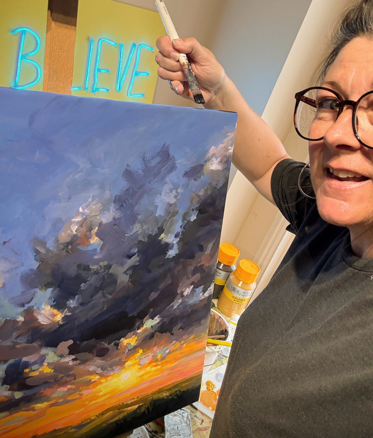 A woman wearing glasses and a dark gray t-shirt is painting a dramatic sunset landscape on a canvas, holding a paintbrush next to an easel with a neon sign that reads 'BELIEVE'.
