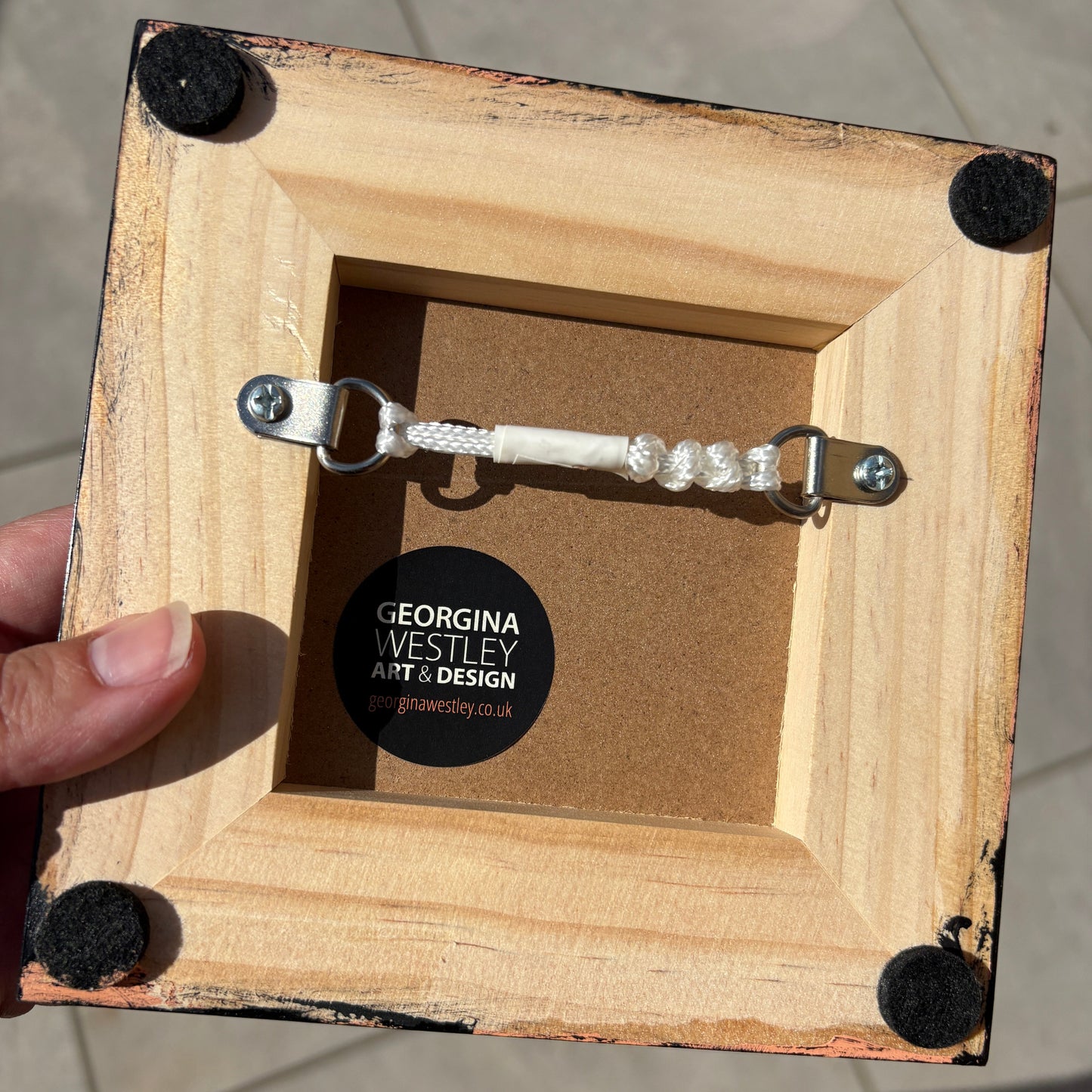 A hand holds a square unfinished wooden picture frame with black corner pads and a hanging wire frame inside. A round sticker on the back reads Georgina Wesley Art & Design.