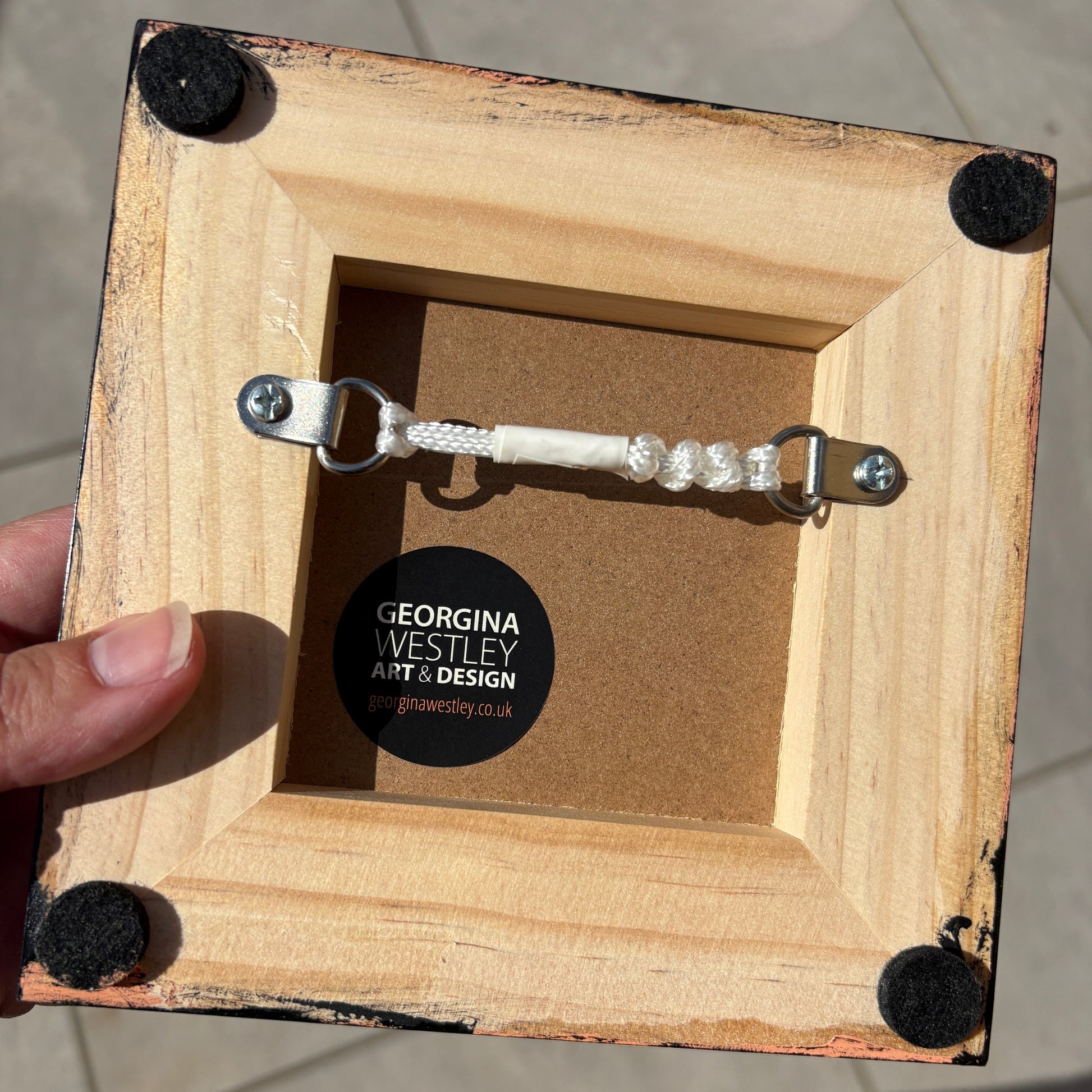A hand holds a square unfinished wooden picture frame with black corner pads and a hanging wire frame inside. A round sticker on the back reads Georgina Wesley Art & Design.