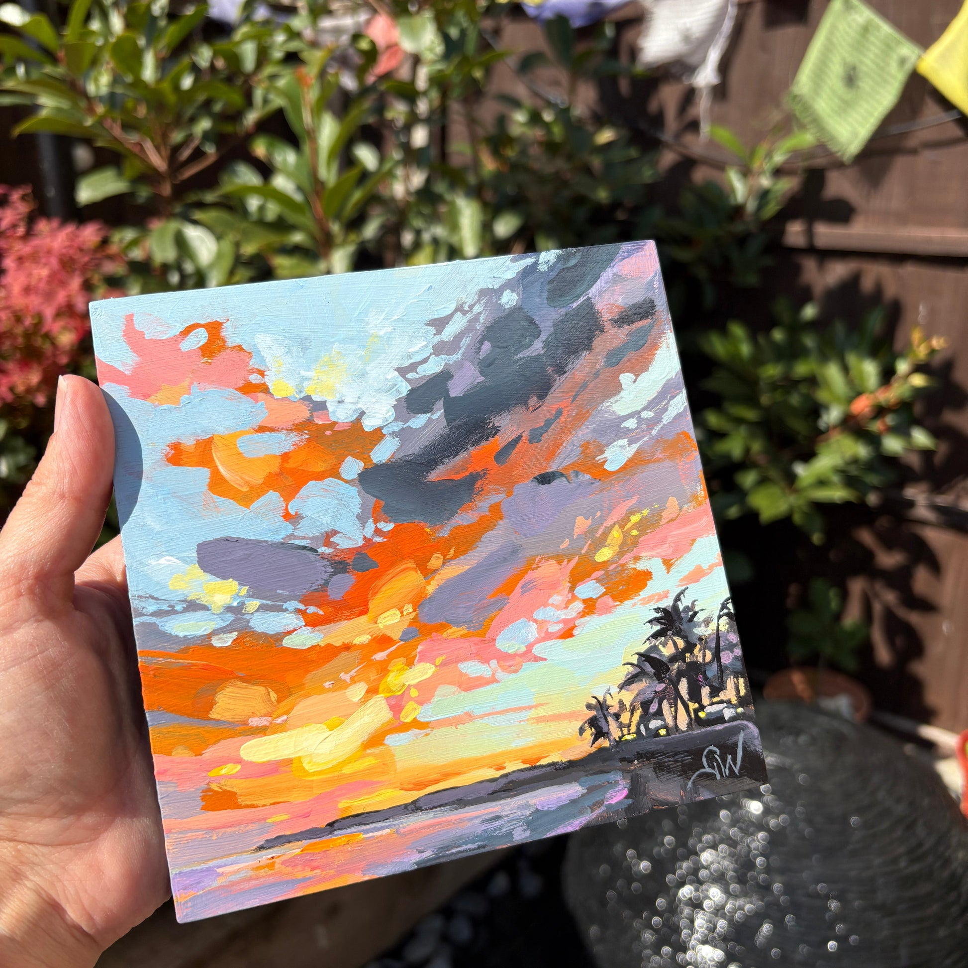 A hand holds a small square canvas painting of a colorful sunset over water with palm trees, photographed outdoors in a garden setting with plants and a fence in the background.