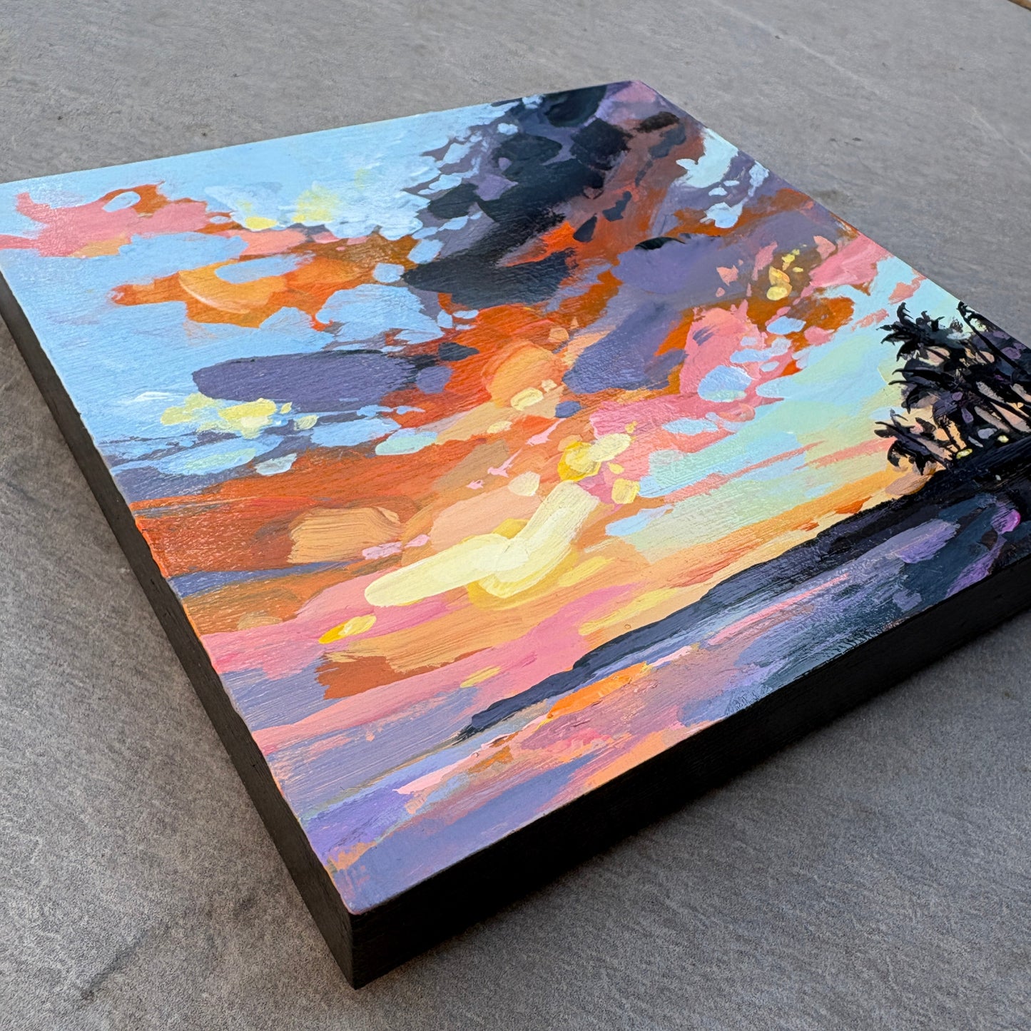 A square canvas painting of an abstract sunset landscape, featuring vibrant brushstrokes of orange, pink, purple, blue, and yellow, displayed on a gray concrete surface.