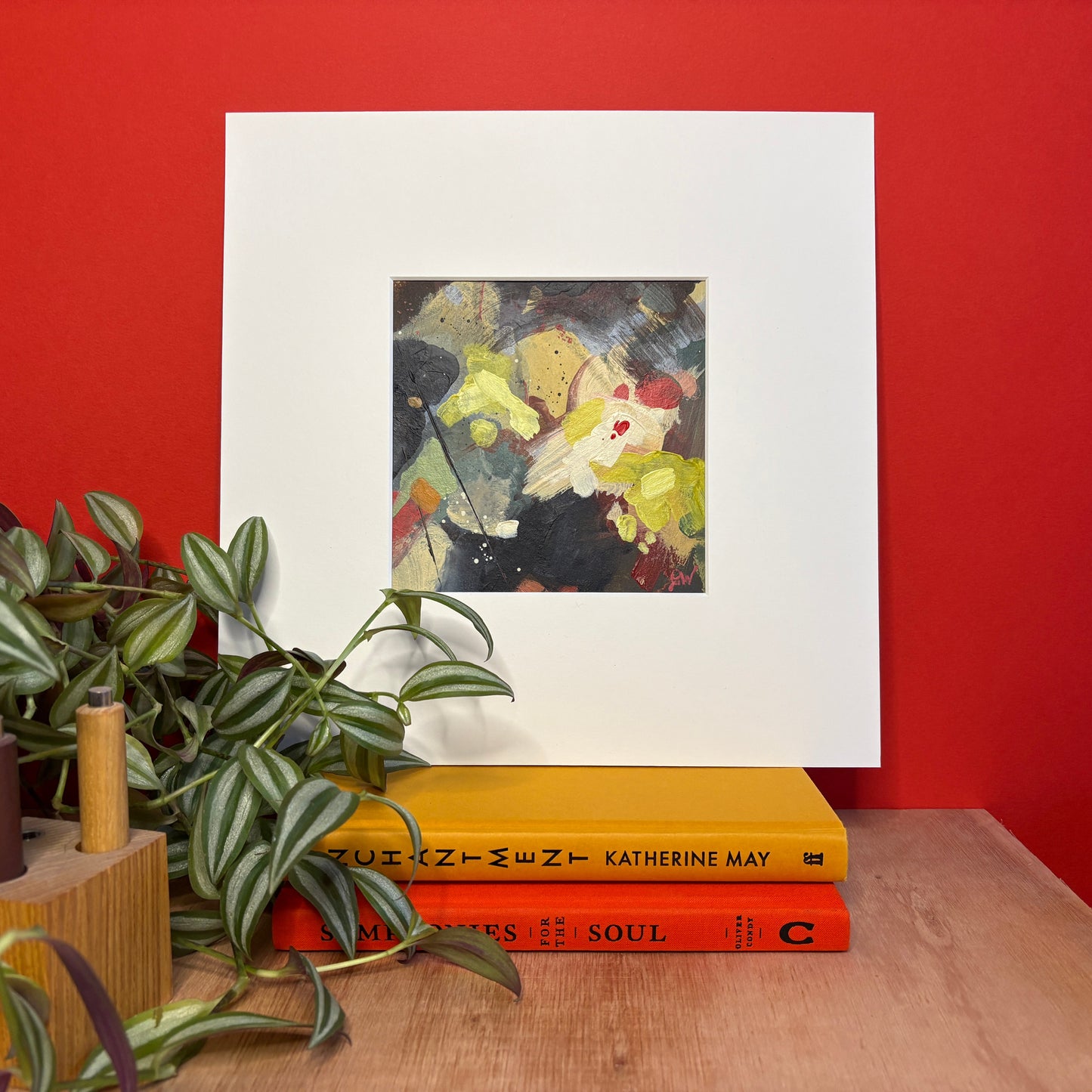 Art inspired by the film Hamnet on a white mount leaning against a red wall with books and plants below.