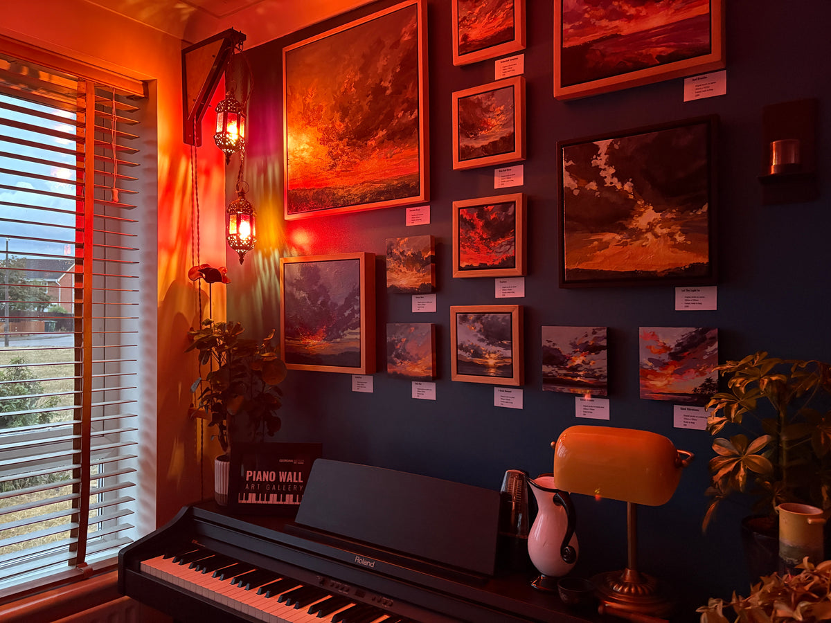 A dark blue wall in a room decorated with multiple framed dramatic landscape paintings glowing under warm orange and red lighting. A black piano sits below the paintings, with a yellow desk lamp, potted plants, and a window with blinds on the left.