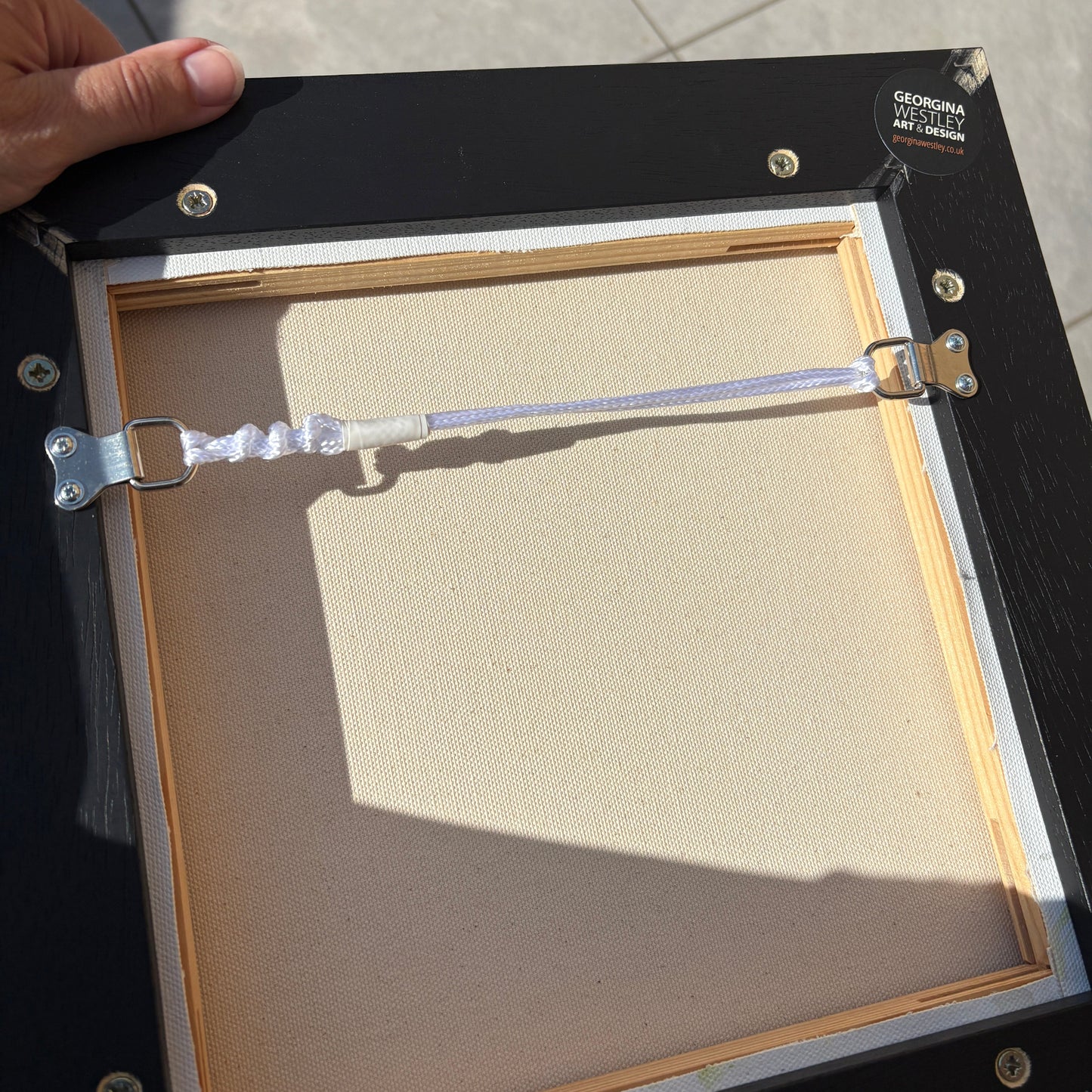 The back of a black picture frame with a wooden mat board, silver metal clips, and a hand holding it on a tiled floor.