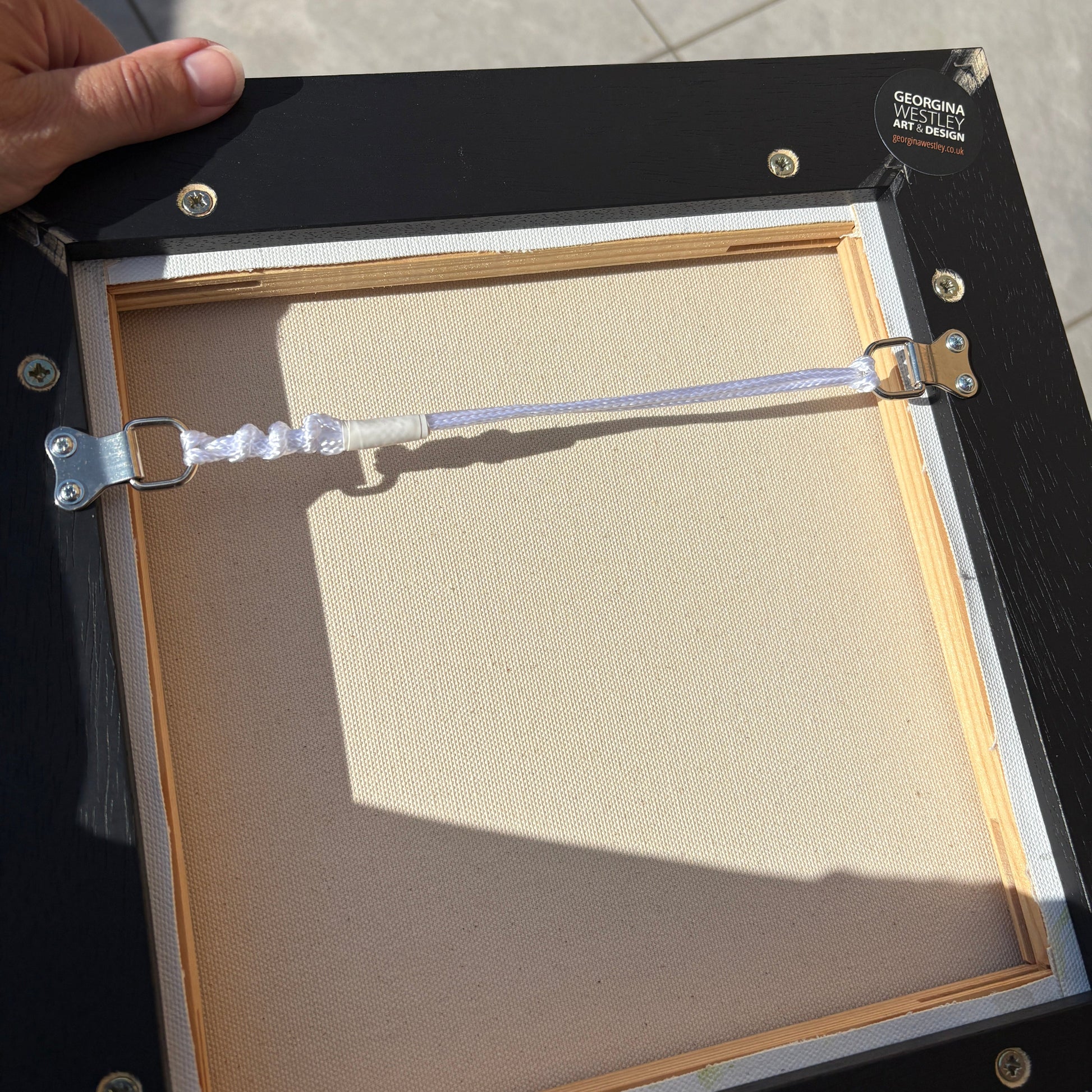 The back of a black picture frame with a wooden mat board, silver metal clips, and a hand holding it on a tiled floor.