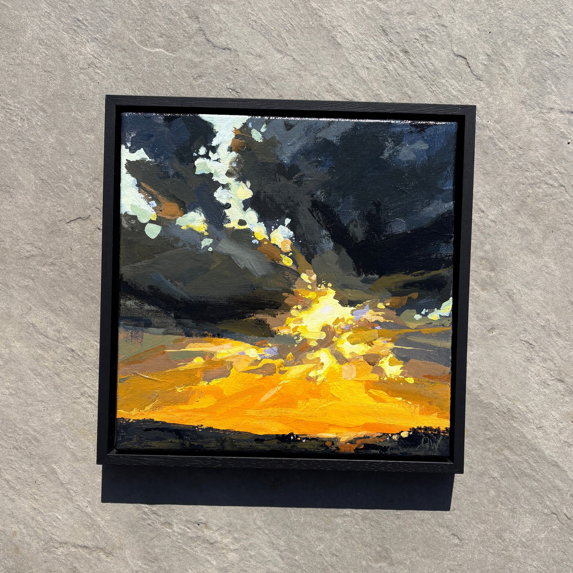 A framed square painting of a dramatic sunset or storm scene, featuring dark gray clouds and a bright yellow-orange glow, mounted on a light stone tile wall.