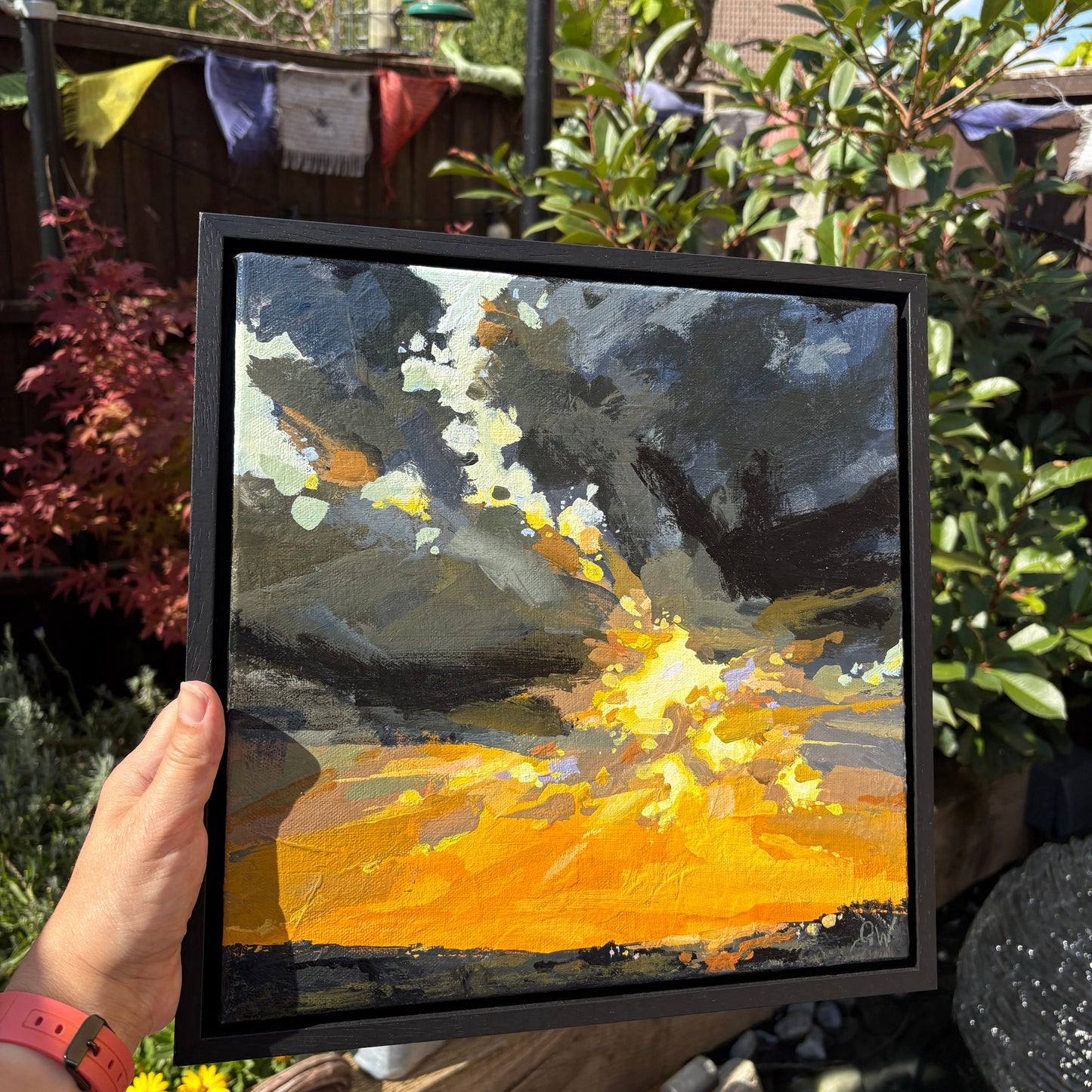 A hand holds a square framed painting outdoors in a garden setting. The painting depicts a dramatic abstract scene with dark gray clouds and a bright glowing yellow-orange ground.