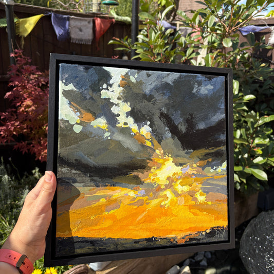 A hand holds a square framed painting outdoors in a garden setting. The painting depicts a dramatic abstract scene with dark gray clouds and a bright glowing yellow-orange ground.