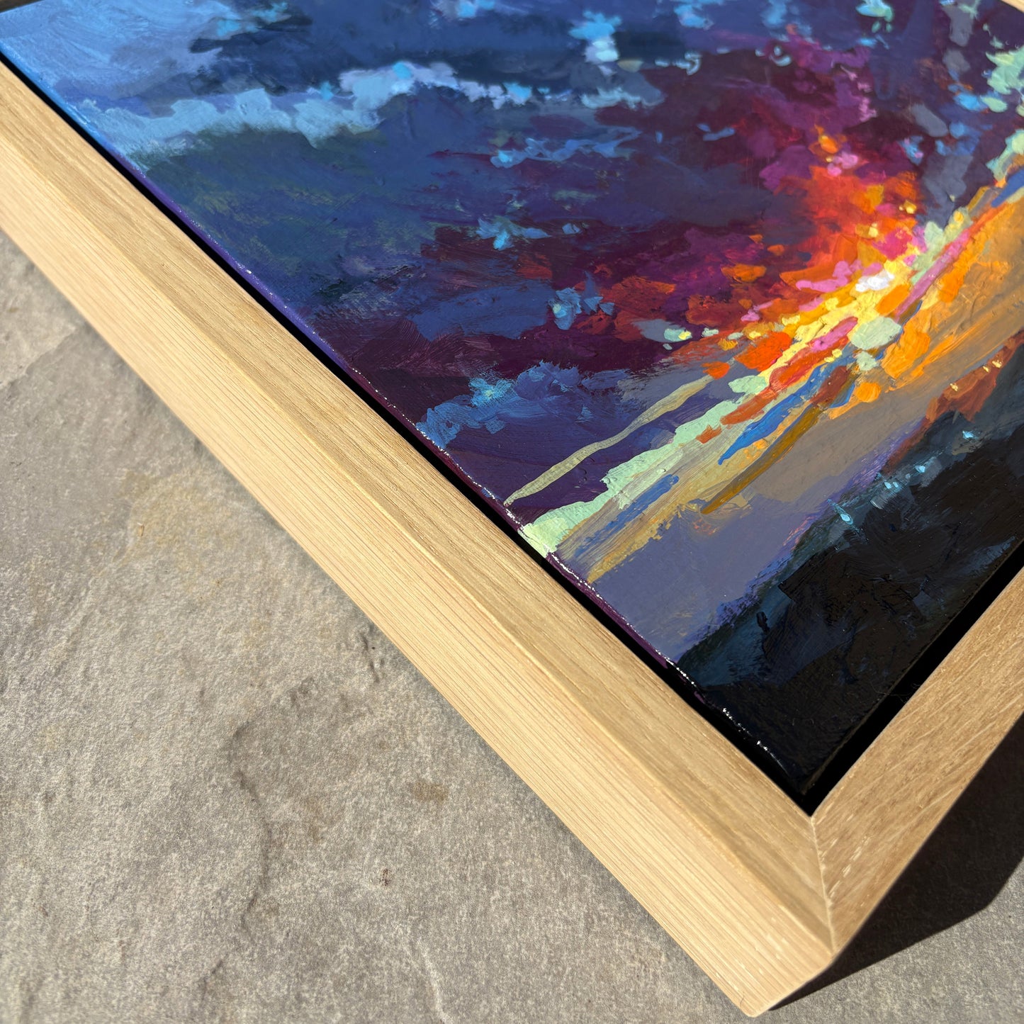 A framed abstract painting featuring a dramatic sky with deep blues, purples, and a bright orange-yellow burst, set in a light natural wood frame and photographed on a gray stone surface.