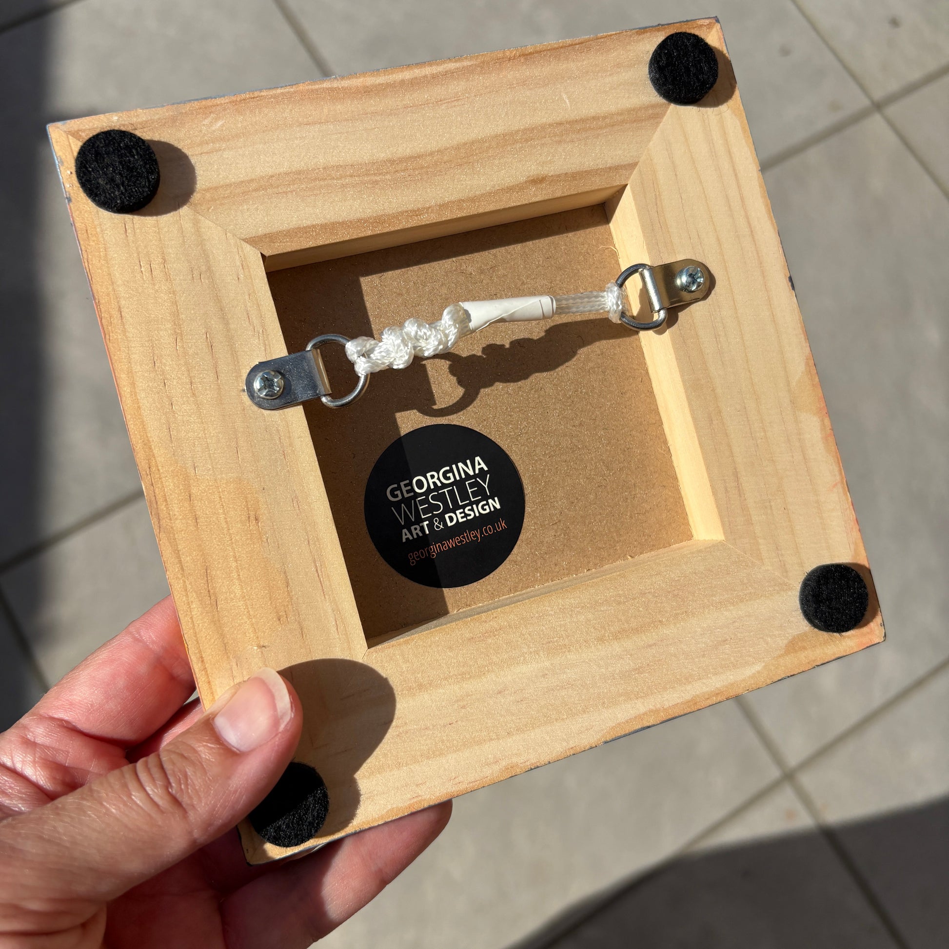 A hand holding a square light wood frame from the back, showing a white rope pull with metal rings, black felt corner protectors, and a circular brand sticker that reads 'Georgina Westley Art Design'.