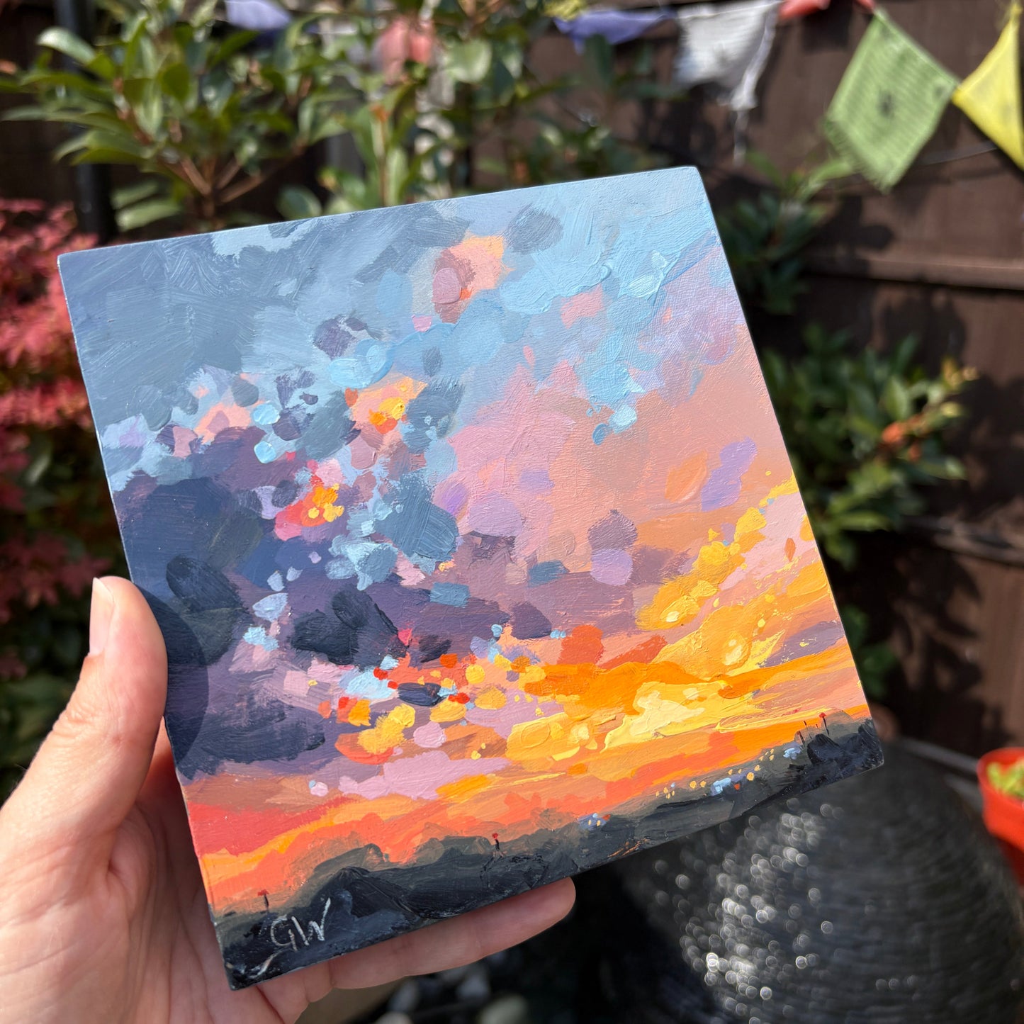 A hand holds a small square canvas painting outdoors in a garden setting. The painting depicts an abstract sunset or sky scene with blue, pink, orange, and yellow tones, and the signature 'GW' is visible in the bottom-left corner.