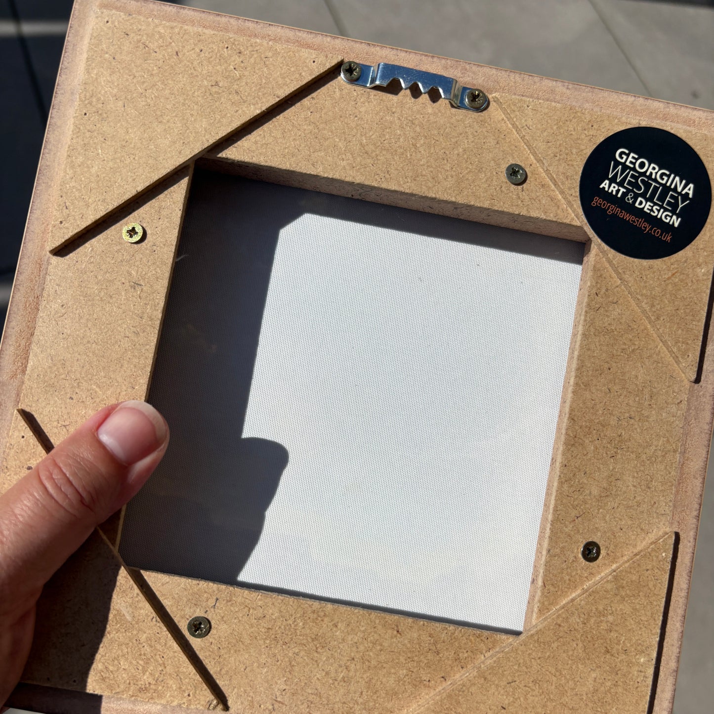 A close-up of a square picture frame backing board made from natural plywood, held by a hand. The back of the frame features pre-drilled screw holes, corner reinforcement strips, and a black circular sticker that reads 'GEORGINA WESTLEY ART & DESIGN'