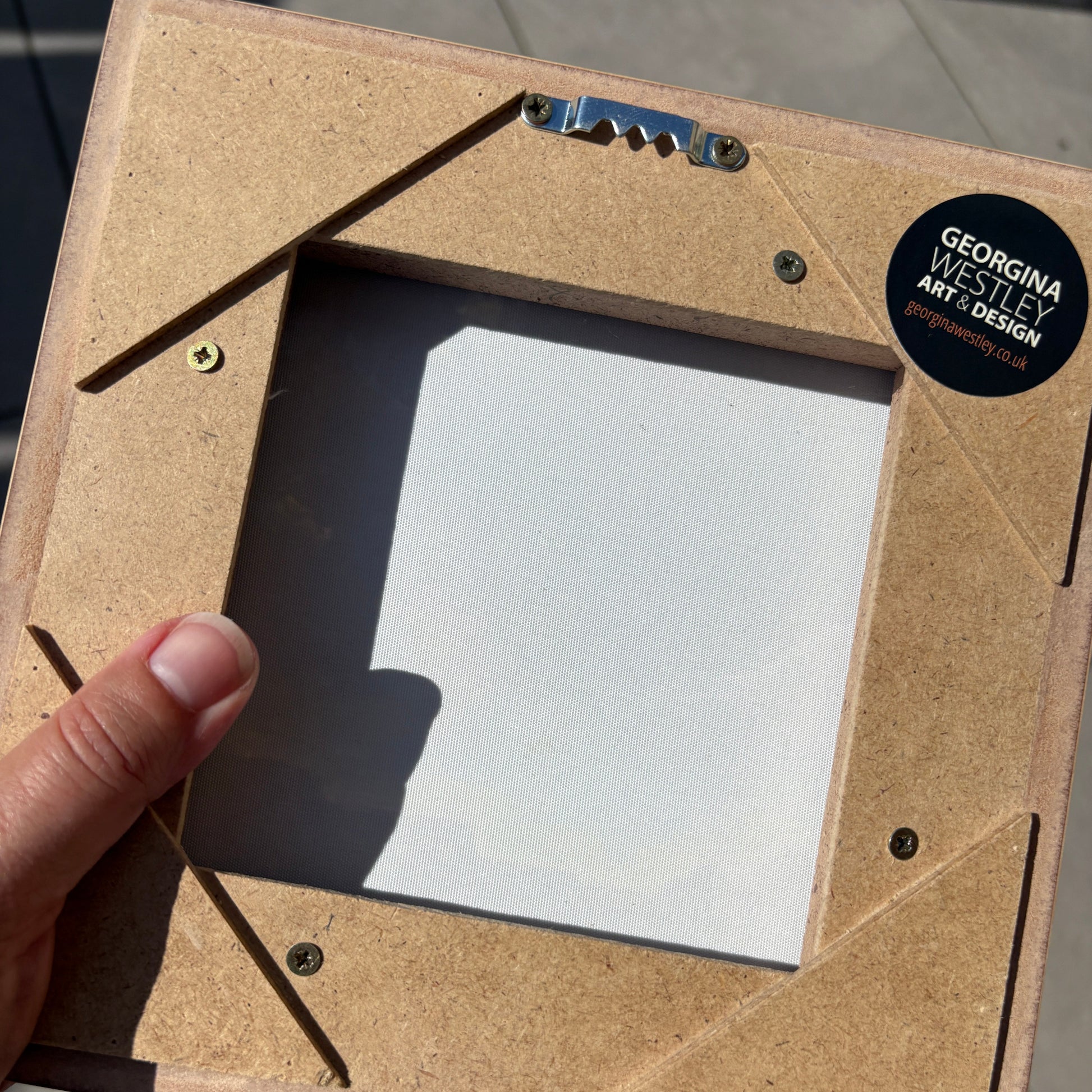 A close-up of a square picture frame backing board made from natural plywood, held by a hand. The back of the frame features pre-drilled screw holes, corner reinforcement strips, and a black circular sticker that reads 'GEORGINA WESTLEY ART & DESIGN'