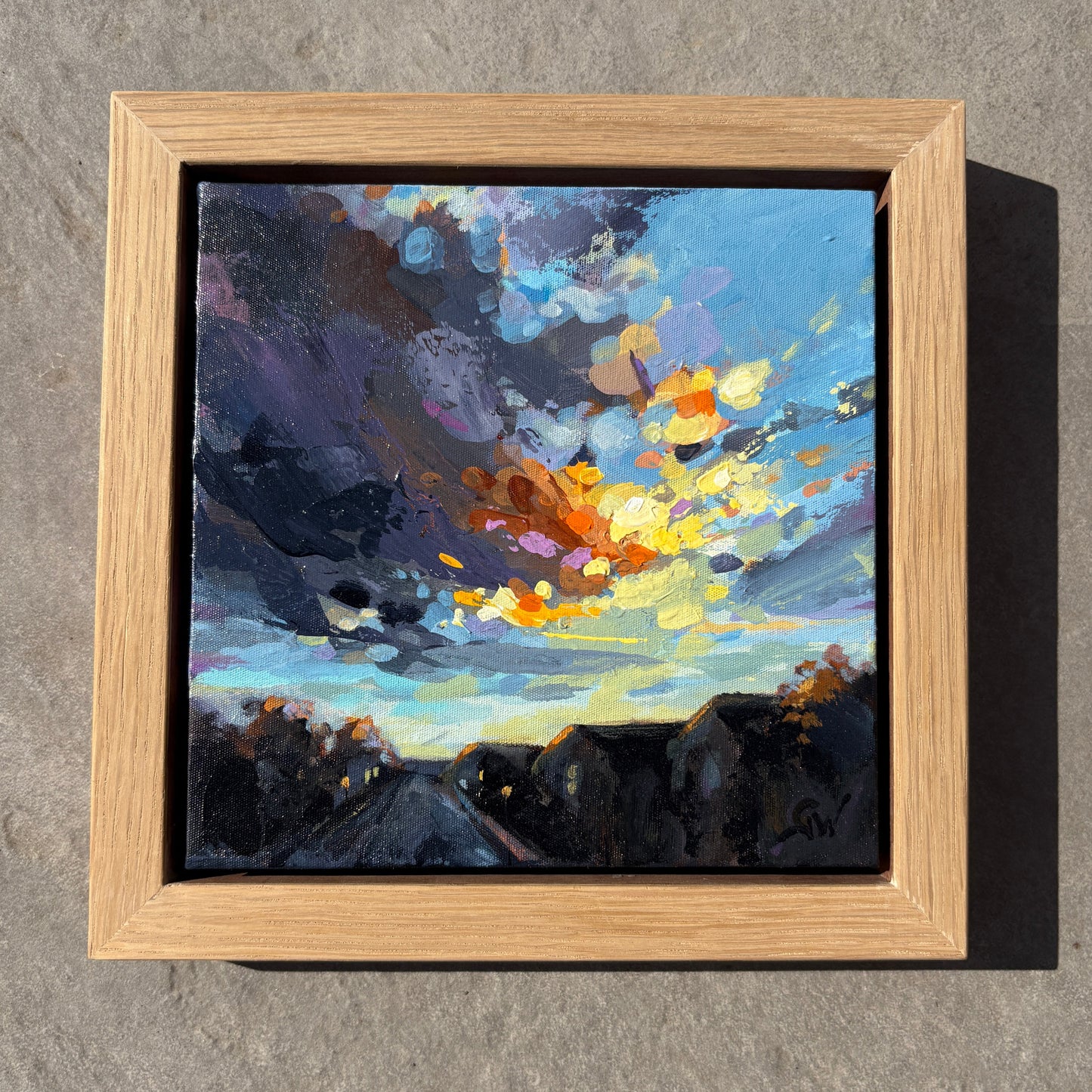 A framed abstract landscape painting featuring a dramatic sky with blue, purple, yellow, and orange brushstrokes above dark silhouetted hills, displayed in a light oak wood frame against a textured gray wall.