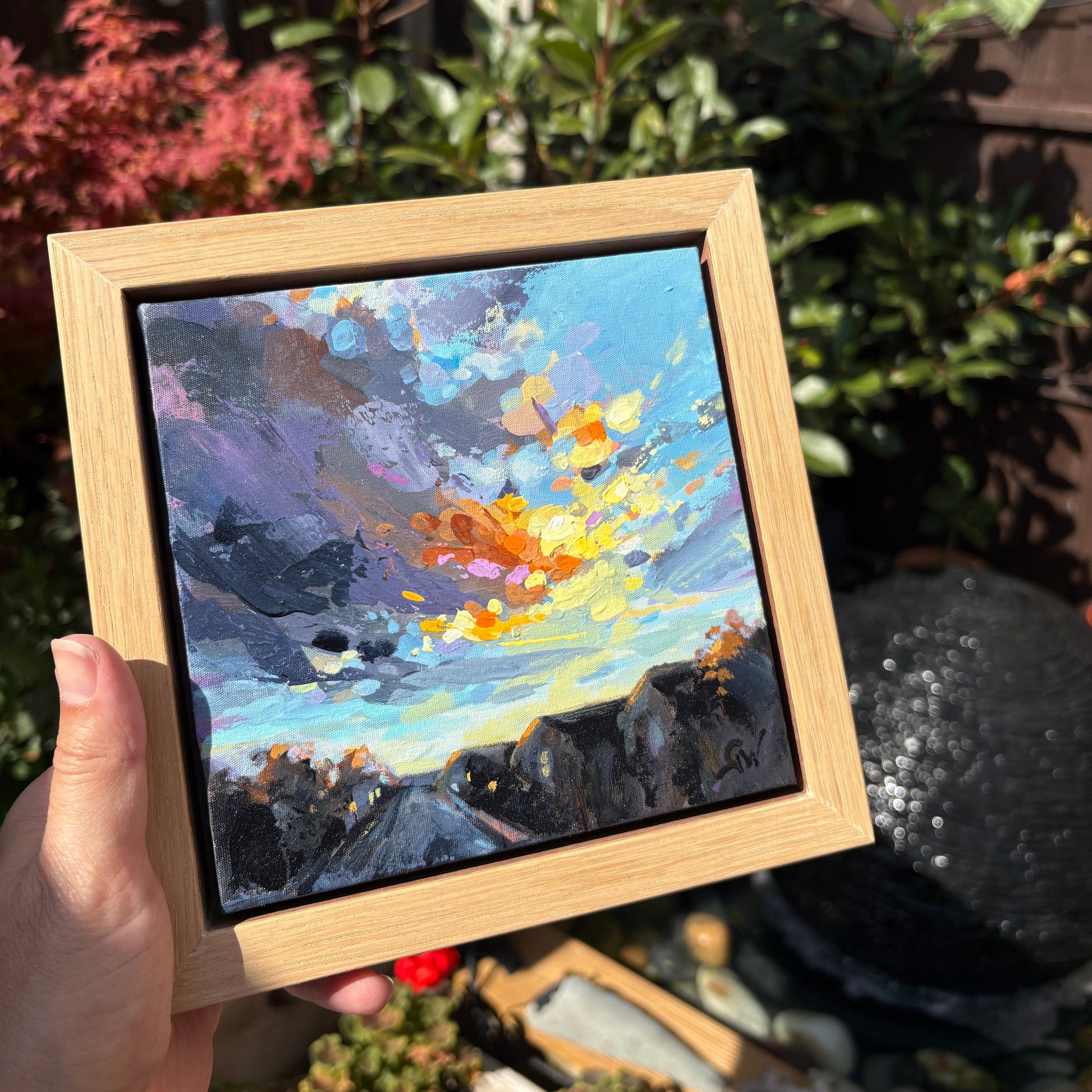 A hand holds a small square framed painting outdoors. The artwork shows an abstract landscape with blue, purple, gray, black, and bright yellow-orange highlights, set in a natural garden background with greenery and a wooden fence.