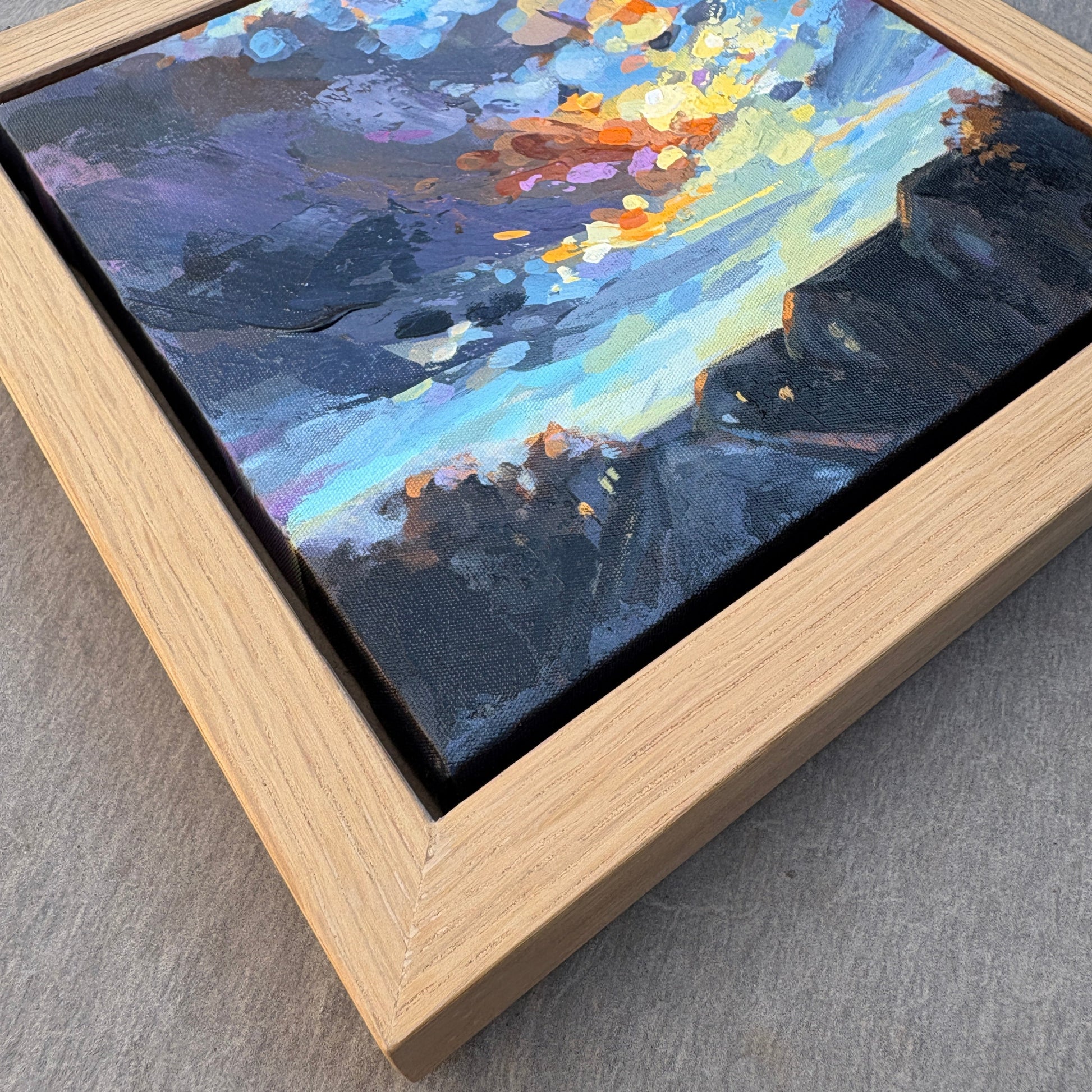 A square framed abstract landscape painting featuring textured brushstrokes of blue, purple, black, yellow, and orange, housed in a light natural wood frame on a gray textured surface.