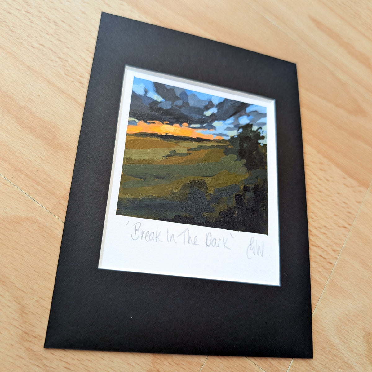 A framed landscape art print titled 'Break In The Dark' by artist Georgina Westley, featuring a textured painting of a field under a stormy sky, matted in black with a white signature band, displayed on a light wood surface.