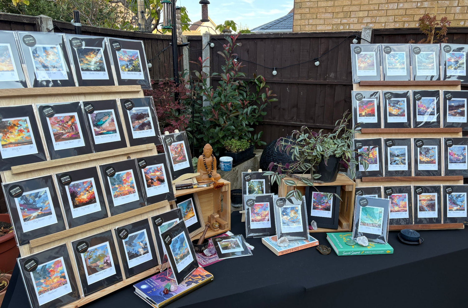 An outdoor market or craft fair display featuring multiple framed art prints and small books arranged on wooden easels and a black-covered table. The prints show colorful landscape scenes, and the background includes a dark wooden fence, plants, and a brick building.