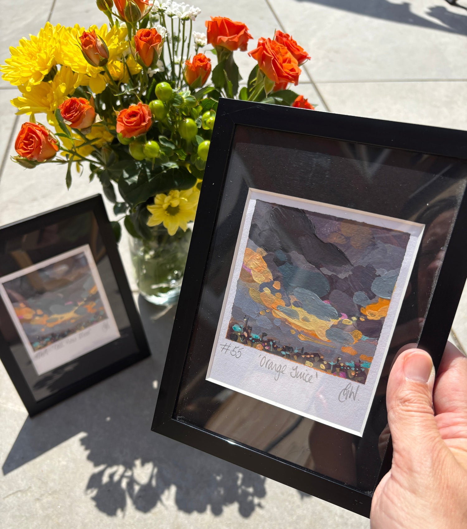 A hand holds a black picture frame containing a colorful landscape art print outdoors on a light concrete surface, with a bouquet of orange and yellow flowers nearby and another framed print below.