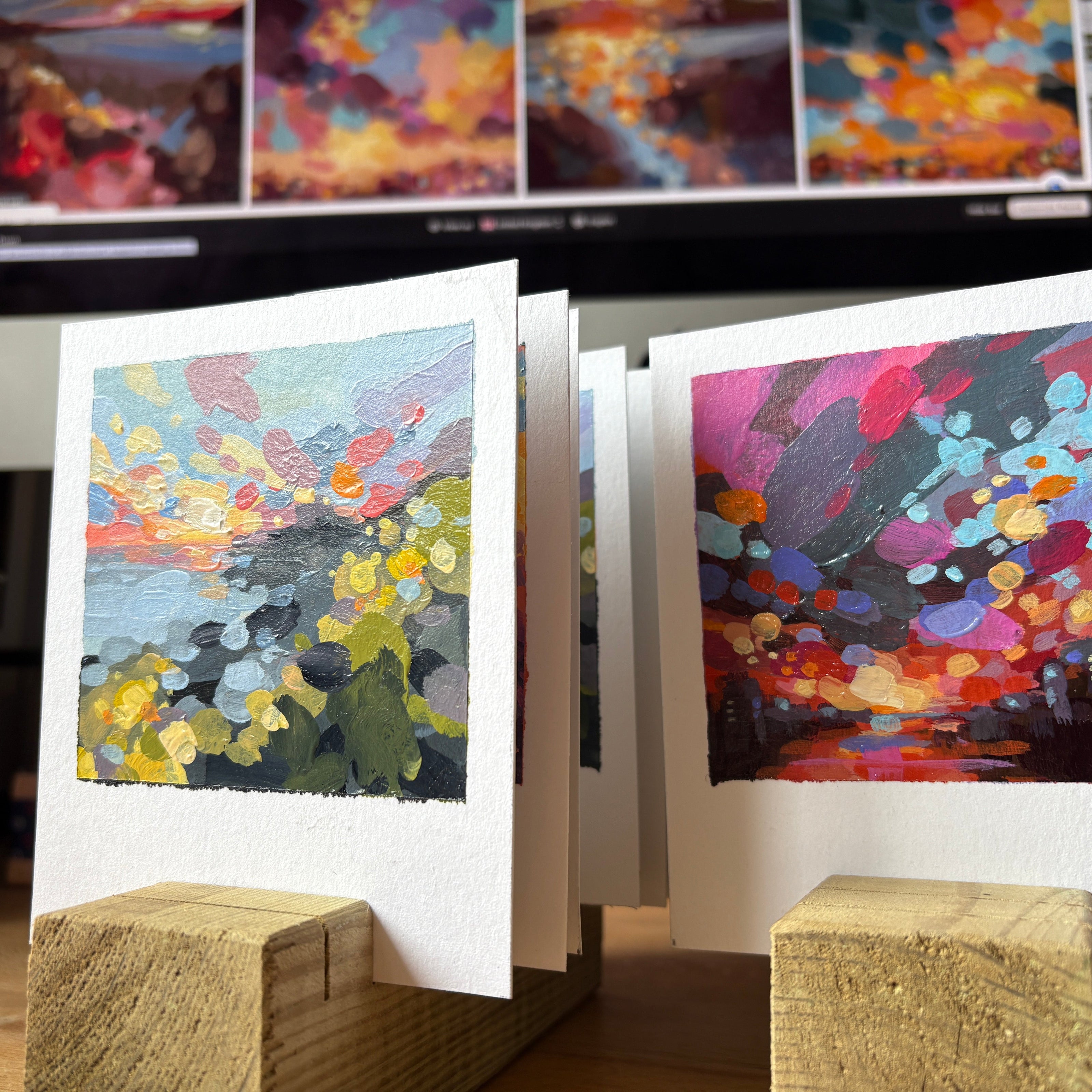 Colorful abstract art prints on stands with a computer screen displaying more artwork in the background.