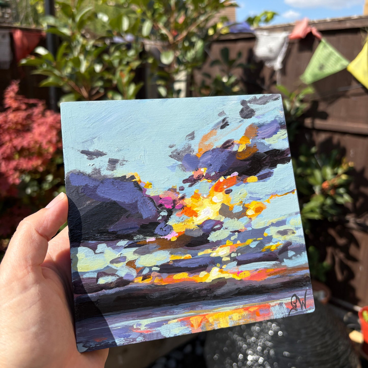 A hand holds a small square canvas painting outdoors in a garden setting. The painting depicts an abstract sunset or storm scene with purple, blue, orange, yellow, and white brushstrokes.