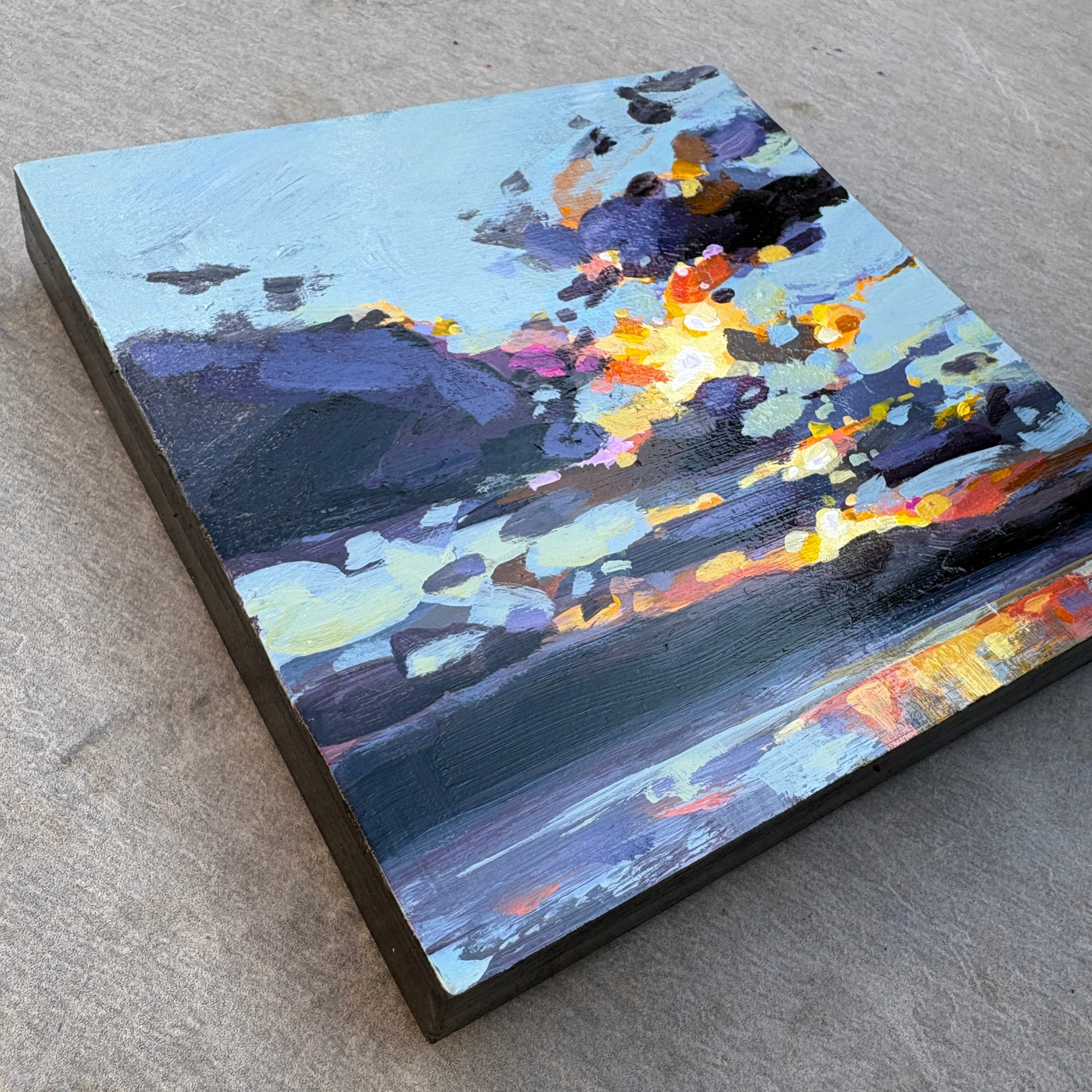A square canvas painting of an abstract landscape or seascape, featuring dark blue, purple, gray, and bright orange-yellow highlights, displayed on a textured gray concrete surface.