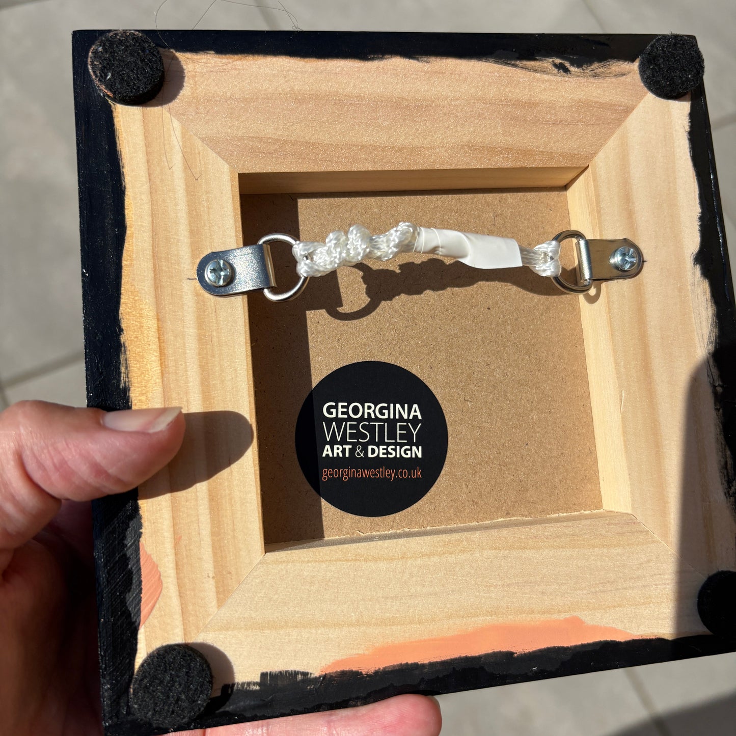 A hand holding the back of a square wooden picture frame. The frame has black painted edges with velvet corners, a silver hanging rod with a white decorative loop, and a round black sticker in the center that reads 'GEORGINA WESTLEY ART & DESIGN'.