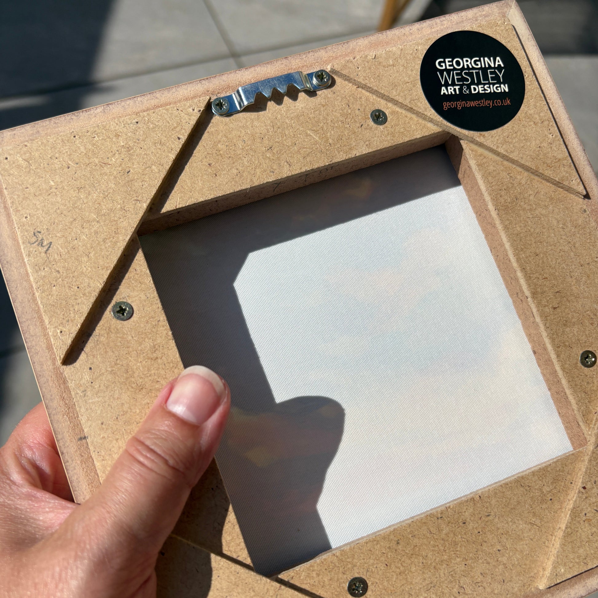 A hand holding a square, angled picture frame made of natural brown cardboard or kraft paper with a clear window. A black circular sticker on the top right reads 'GEORGINA WESTLEY ART & DESIGN'.