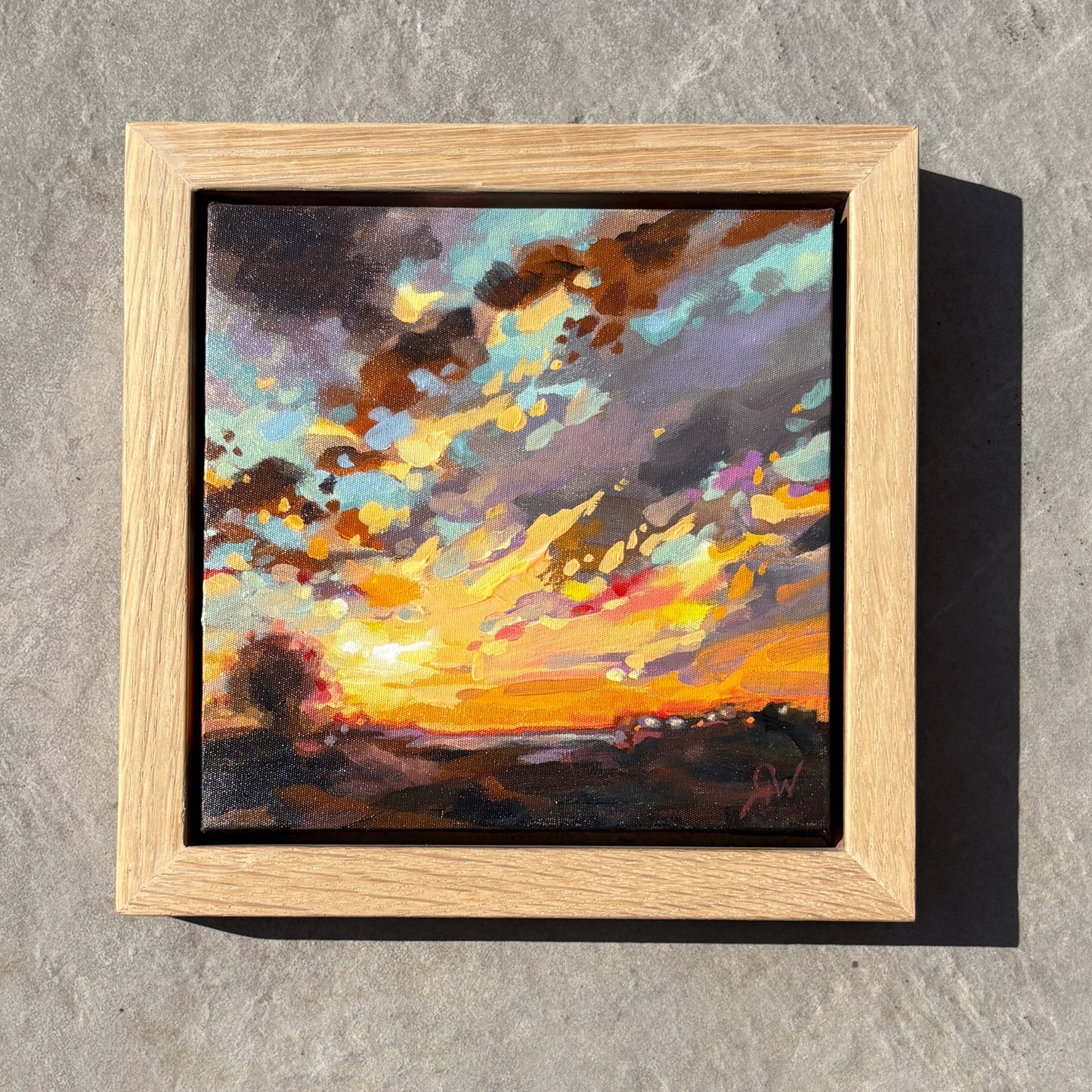 A square framed abstract painting featuring vibrant orange, yellow, and blue brushstrokes, set in a light wood frame on a gray stone surface.